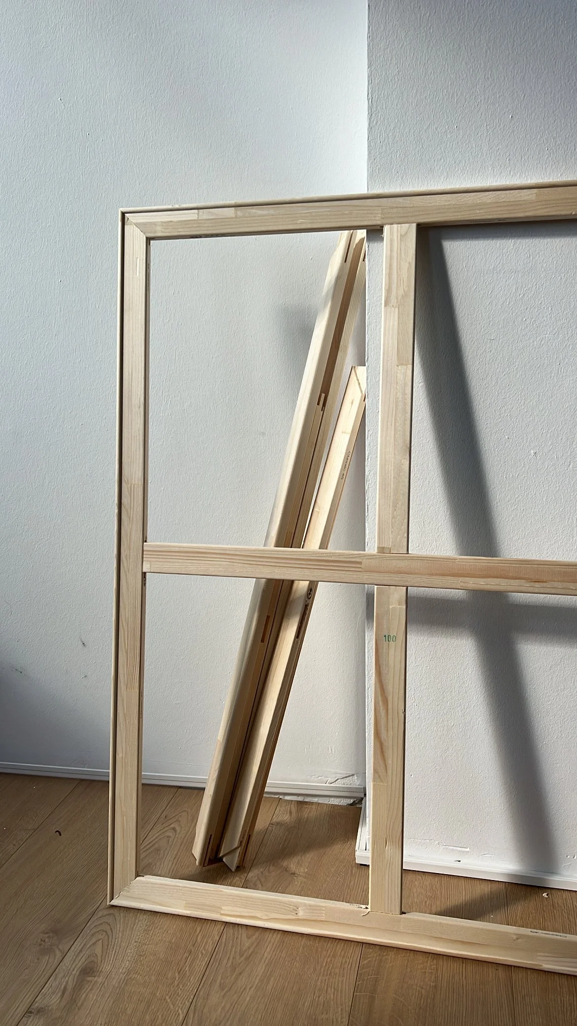 A wooden frame of a miniature or small structure leaning against a white wall with wooden flooring, with some wooden boards inside the frame.