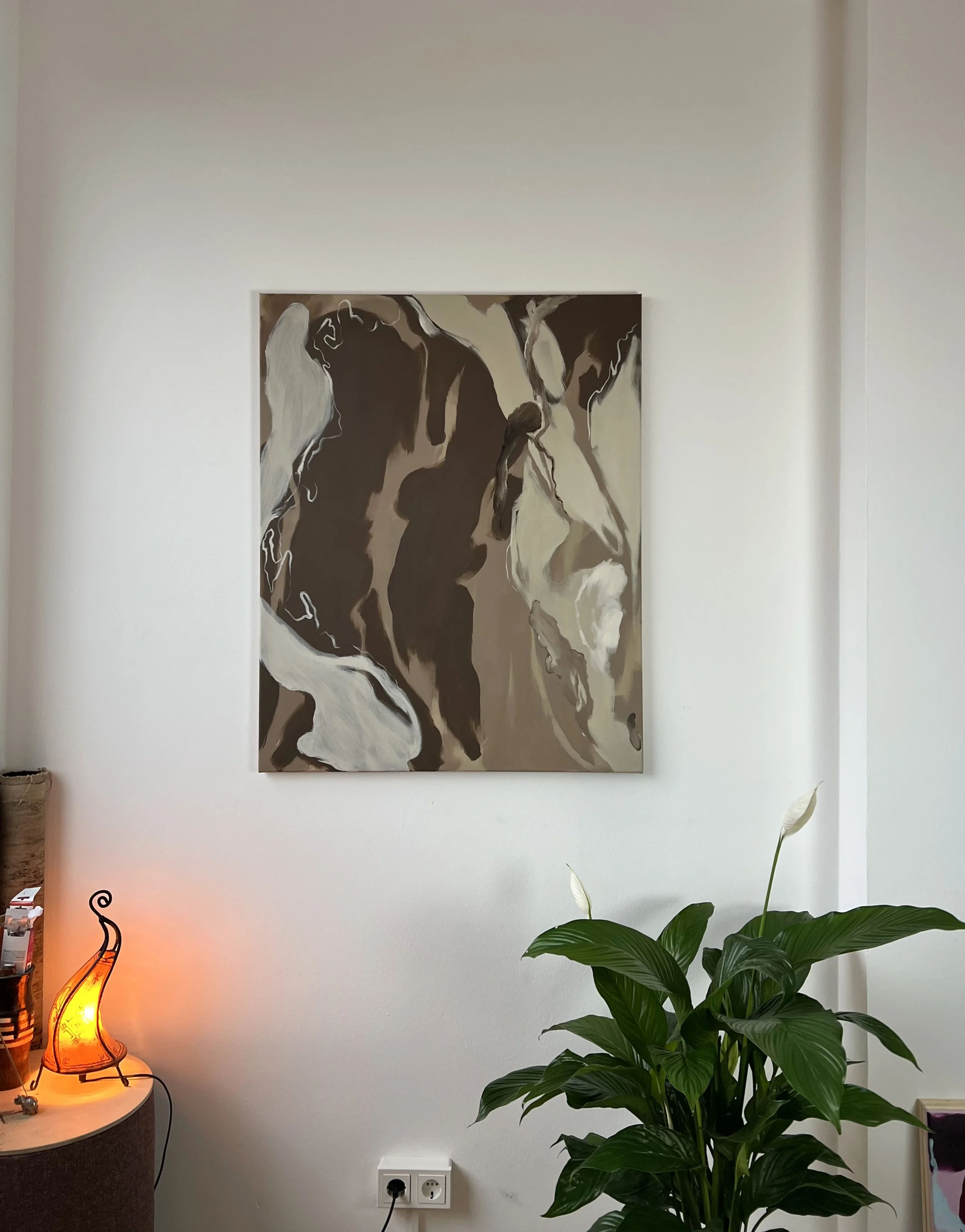 A modern living room corner featuring a large potted green plant with broad leaves, above a white baseboard, a painting of two abstract figures on the wall, and a small side table with a decorative orange lamp, a small alarm clock, and some items. Th