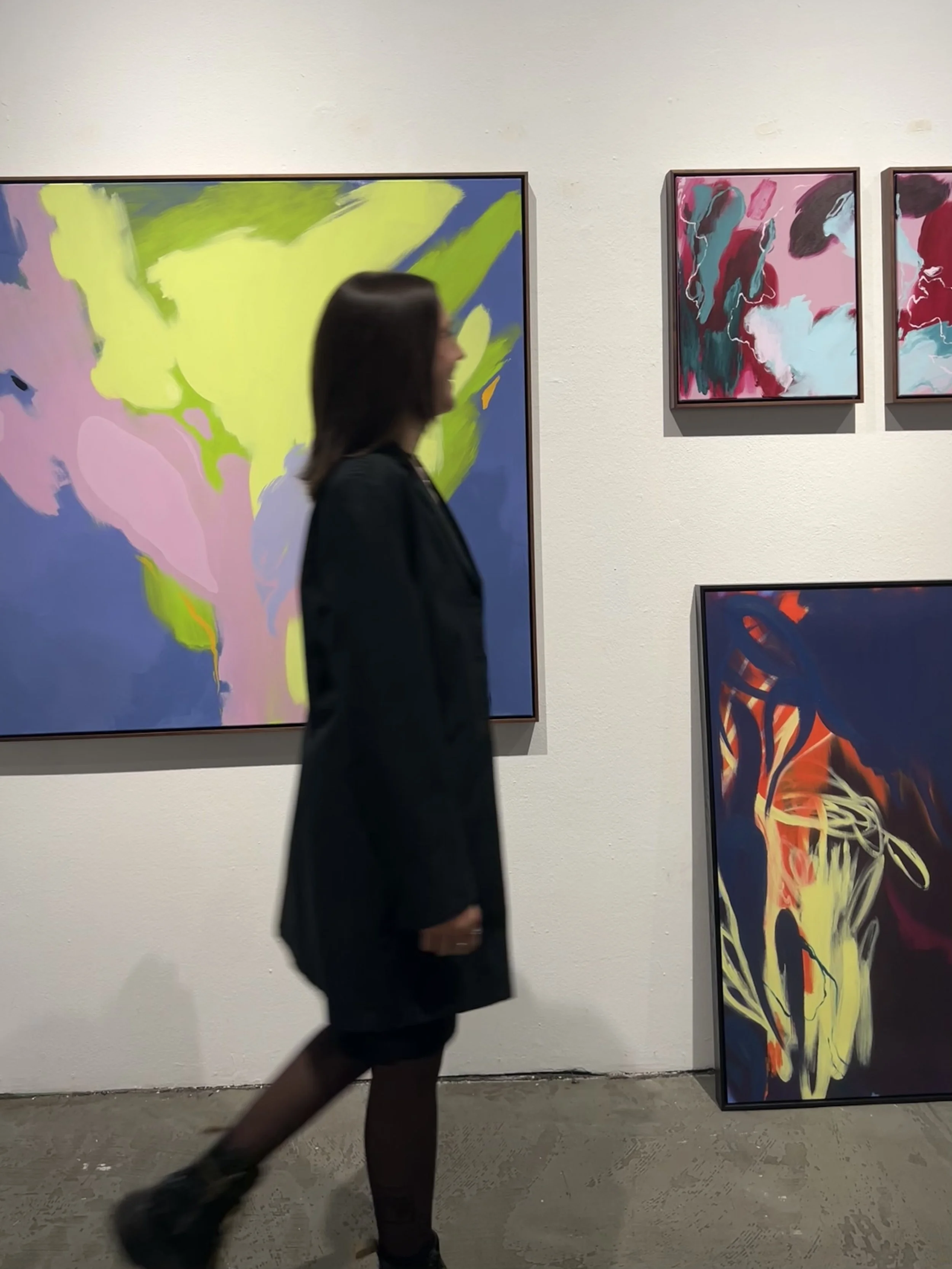 A woman dressed in black walking past colorful abstract paintings displayed on a white gallery wall.