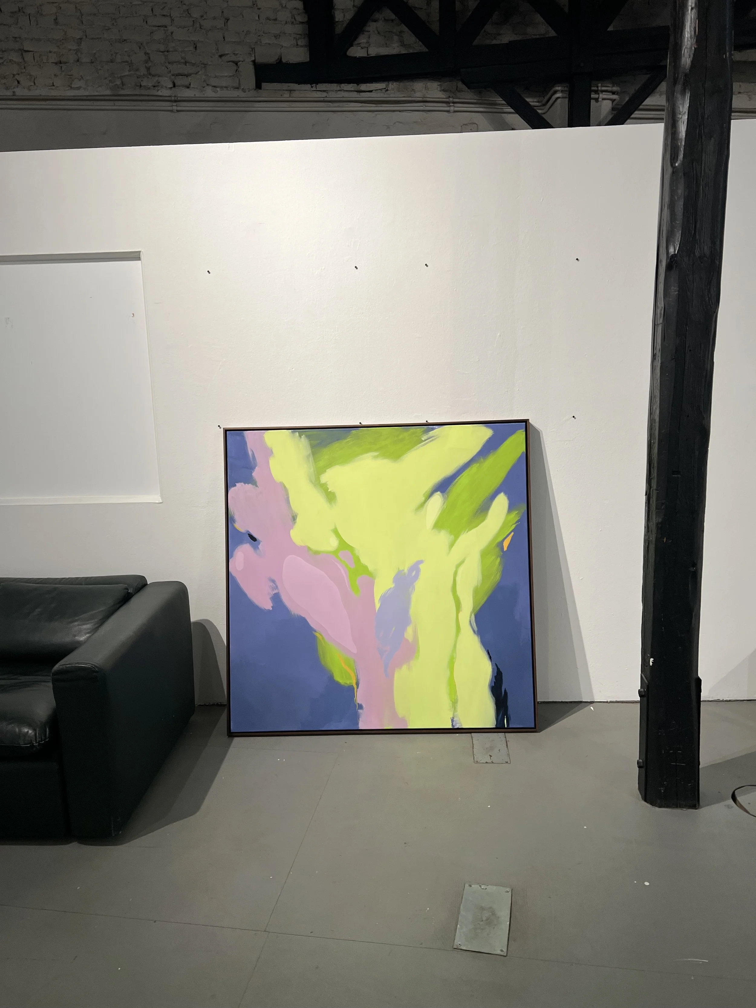 An abstract colorful painting with shades of green, pink, purple, and dark blue leaning against a white wall in an industrial-style room with exposed brick and wooden beams.