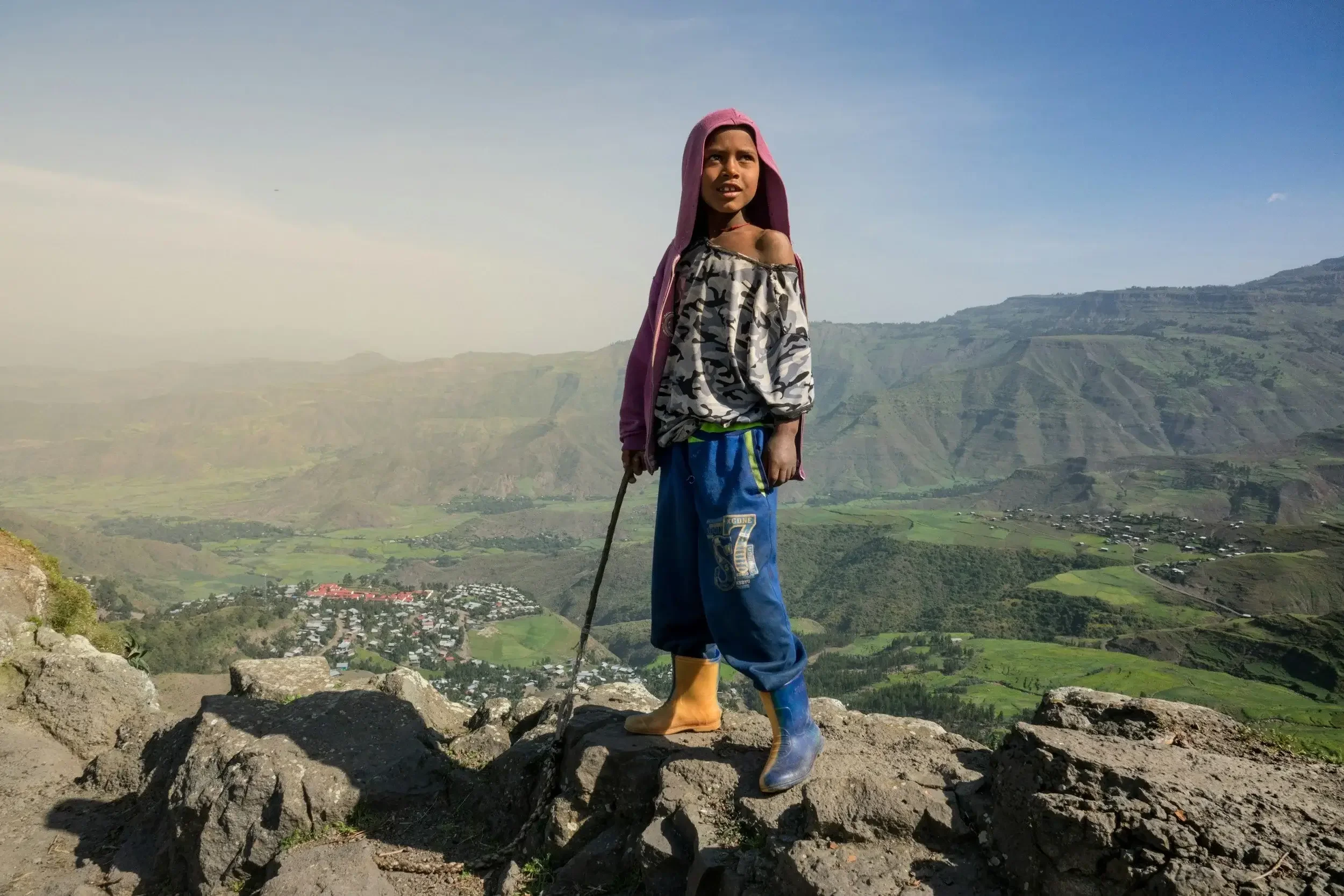 Young boy standing on rocks on a hillside overlooking a valley with fields and scattered buildings, wearing a pink hoodie, off-shoulder shirt, blue pants, yellow boots, and holding a stick, with a mountain landscape in the background.