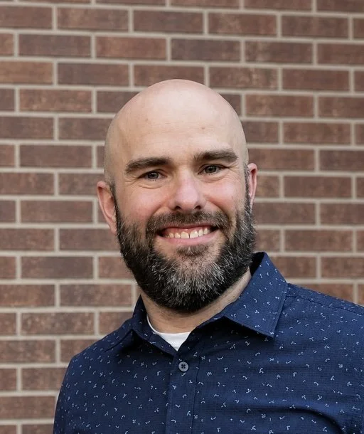 A man with a bald head and a beard, smiling, wearing a blue patterned shirt, standing in front of a brick wall.