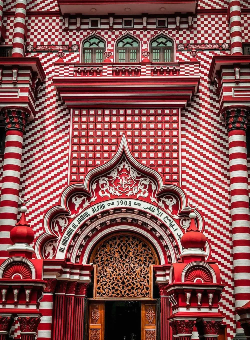 Close-up of a richly decorated building facade with red and white striped patterns, ornate archway, and Arabic script. The entrance features an intricately designed metal gate with wooden doors. The architecture includes arches, columns, and decorative elements typical of Islamic or Moorish styles.