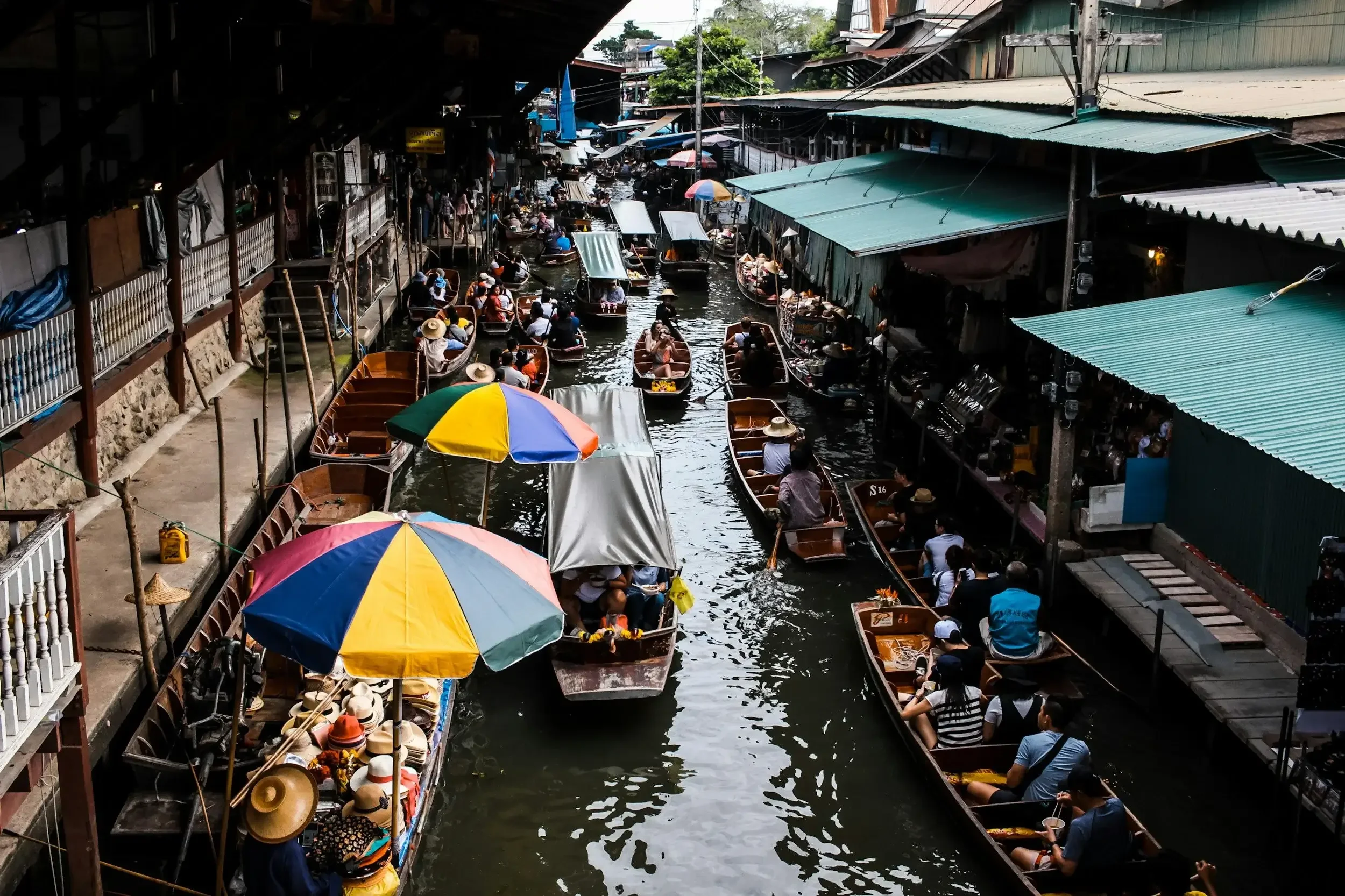 A crowded floating market with boats filled with people and stalls along a canal. Some boats are shaded with umbrellas and the market is surrounded by buildings with green roofs and wooden structures.