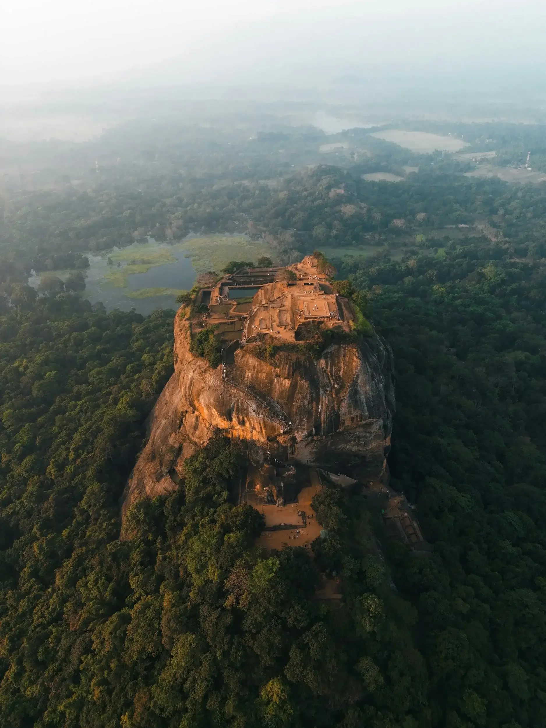 Aerial view of Sigiriya Rock Fortress, an ancient palace and rock formation in Sri Lanka, surrounded by lush green forest and farmland, with hazy sky in the background.