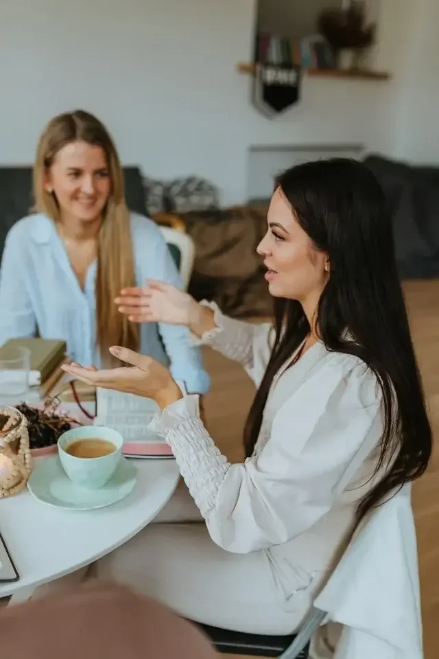 Two women sitting at a table, engaged in a conversation. One woman with long black hair is gesturing with her hand, and the other woman with blonde hair is smiling. The table has a cup of coffee, a basket, and some books or notebooks.
