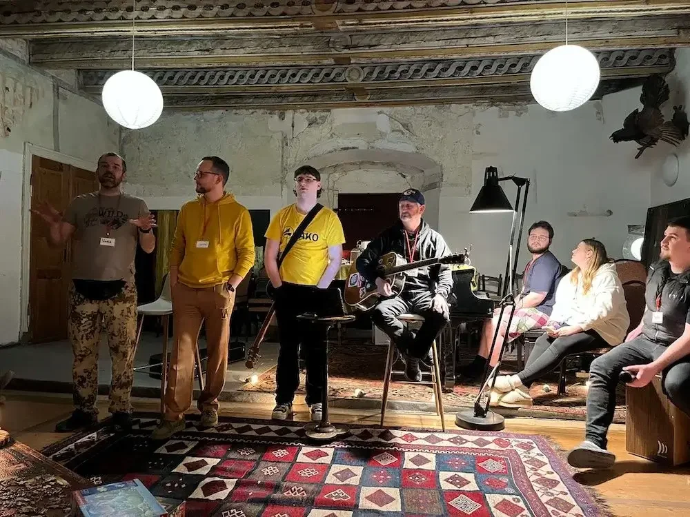 Group of people at a casual gathering, with some sitting and some standing, in a rustic room with a carpet, a guitar, and paper lanterns hanging from the ceiling.