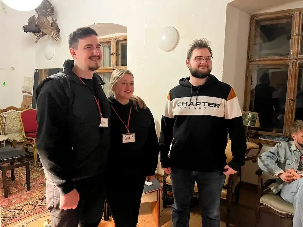 Three young adults with name tags standing together in a warmly lit room, smiling, with another person sitting nearby, and decorative items like a taxidermy bird on the wall and vintage furniture in the background.