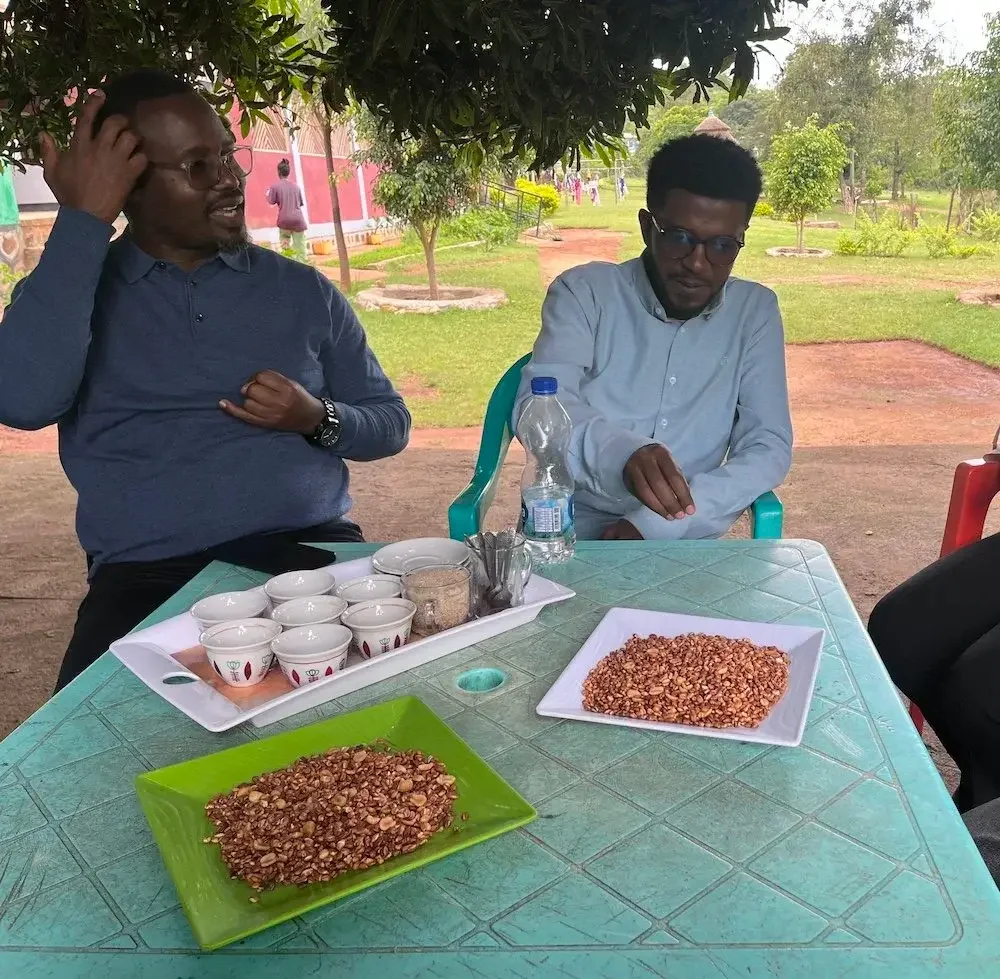Two men sitting at a table outdoors under a tree with trays of snacks, cups, and a bottle of water.