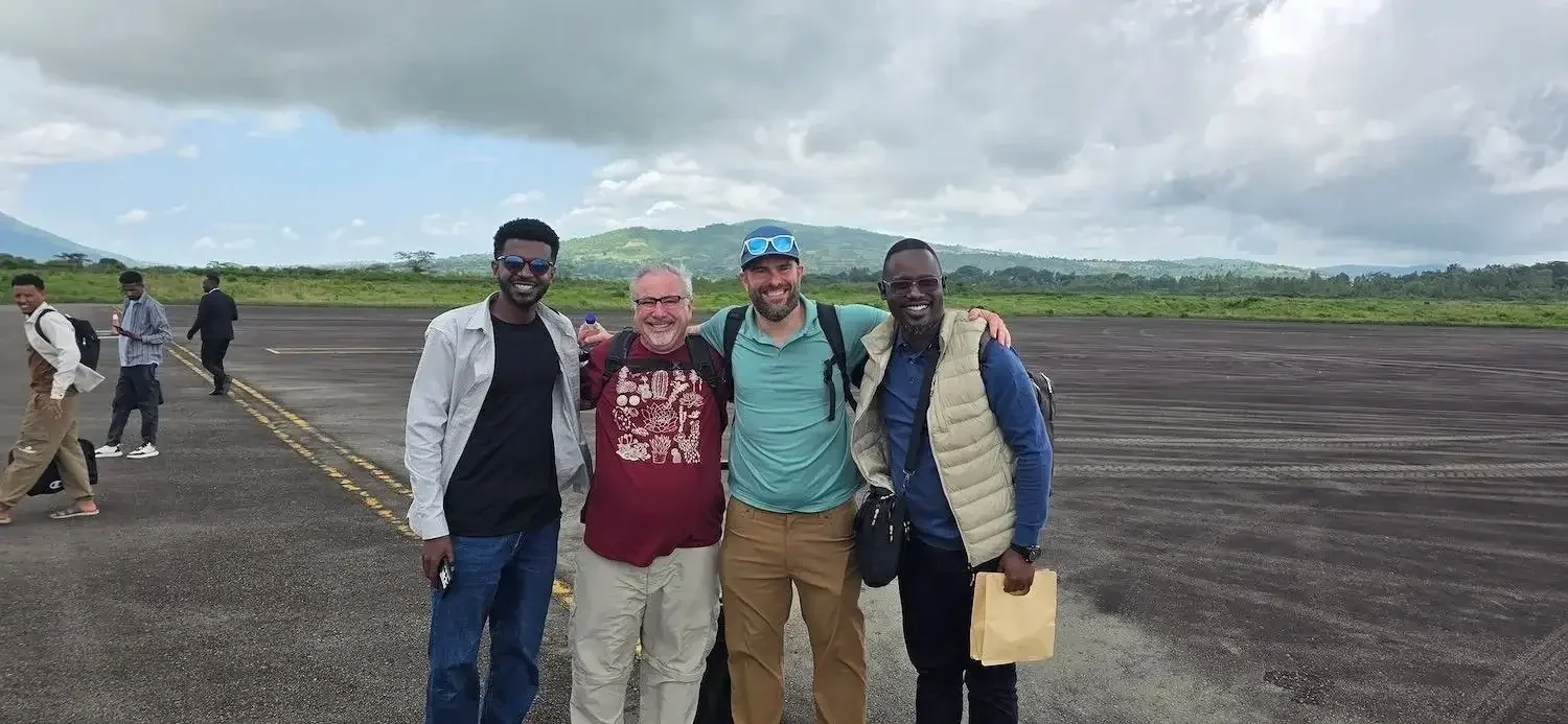 Four men standing together on an airport tarmac, smiling and posing for a photo, with two other people in the background, a landscape with green hills and partly cloudy sky.