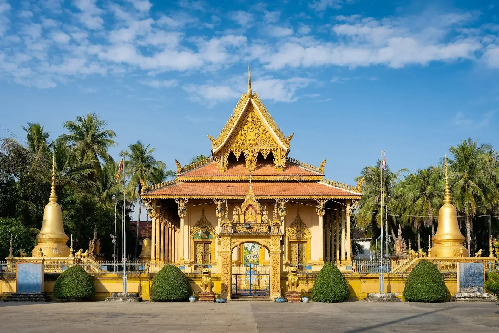A traditional Thai temple with ornate gold details, surrounded by green palm trees and statues, under a blue sky with scattered clouds.