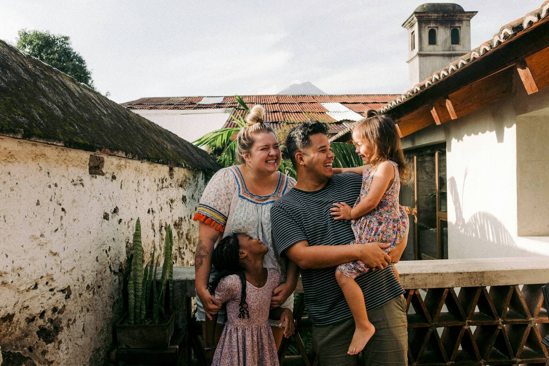 A diverse group of five people, including children and adults, smiling and talking outdoors on a sunny day, with a house, plants, and a mountain in the background.