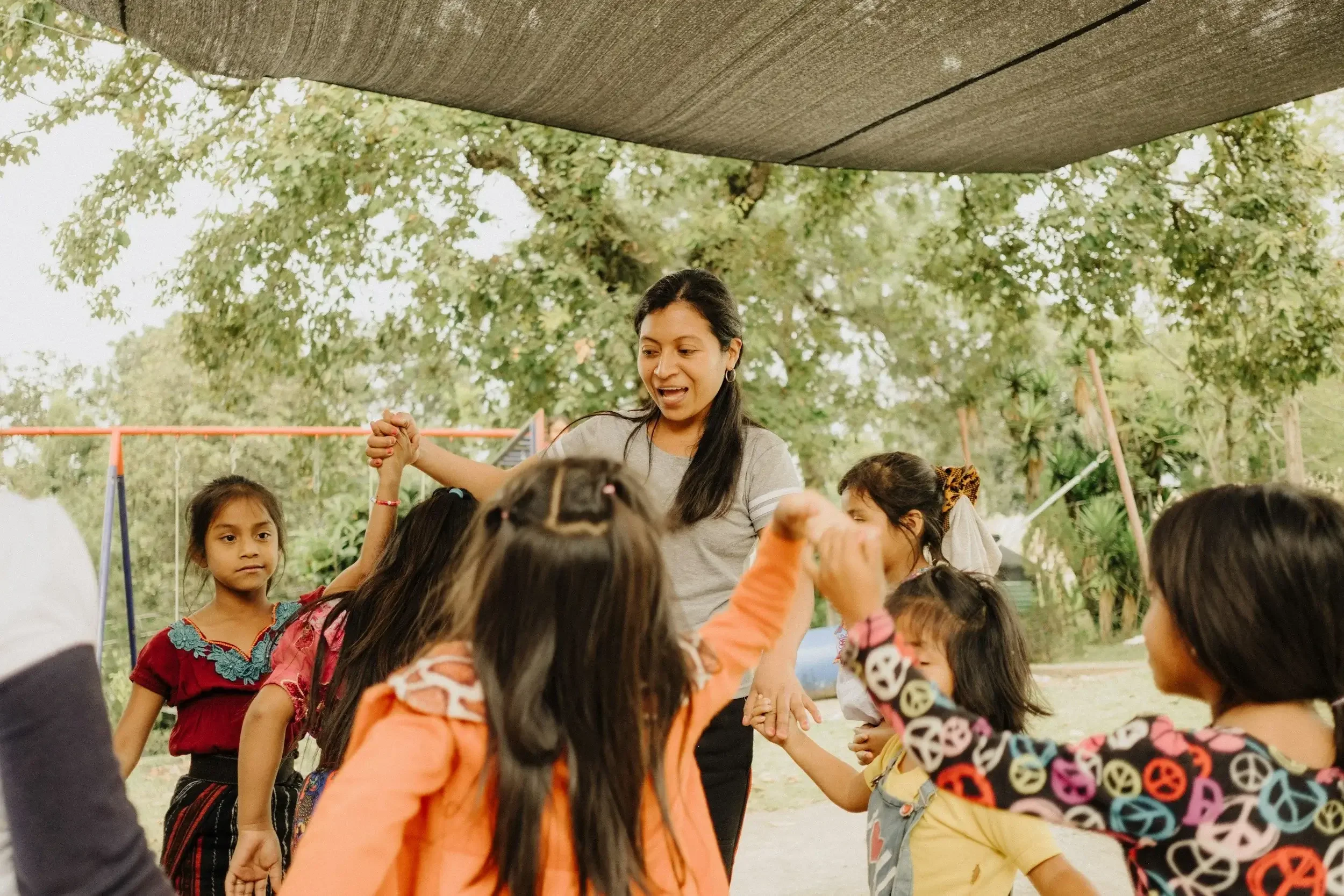 A group of young children holding hands in a circle outdoors with a woman in the center, under a canopy with trees in the background.