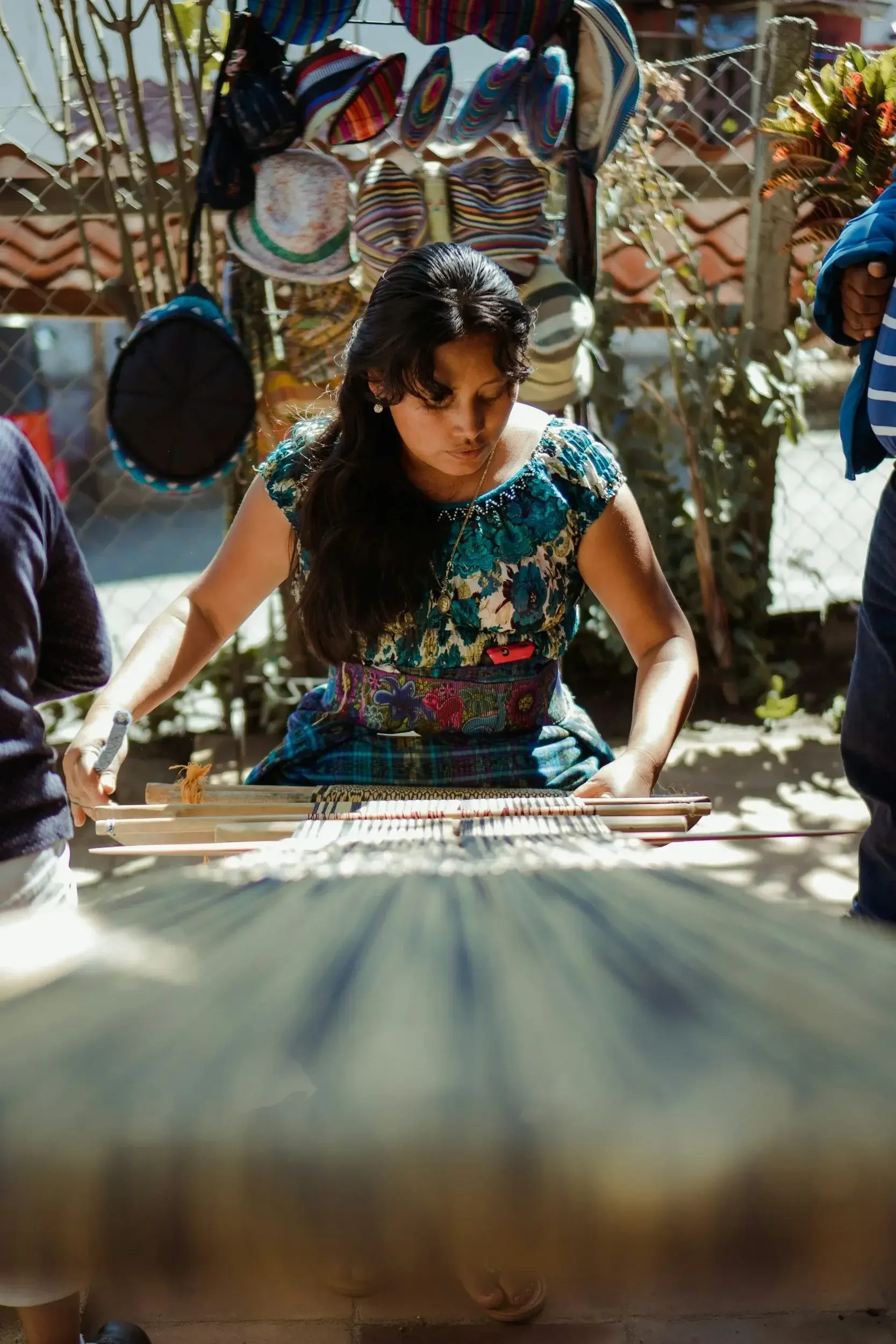 A woman dressed in traditional clothing is weaving on a loom outdoors. Behind her, a chain-link fence displays colorful bags and hats made from striped and patterned fabric. The setting appears to be in a sunny, possibly rural area with trees and a tiled roof in the background.