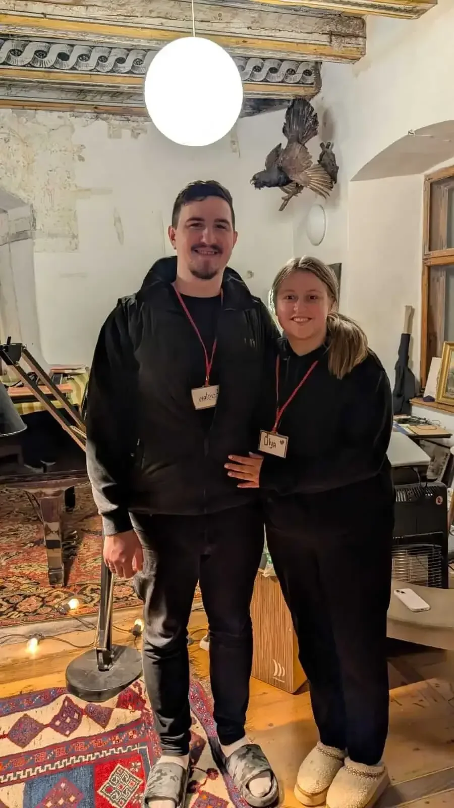 A man and a girl standing together indoors, smiling, wearing black hoodies with name tags, in a cozy room with wooden beams, a hanging light, a mounted bird, and various framed pictures and furniture.
