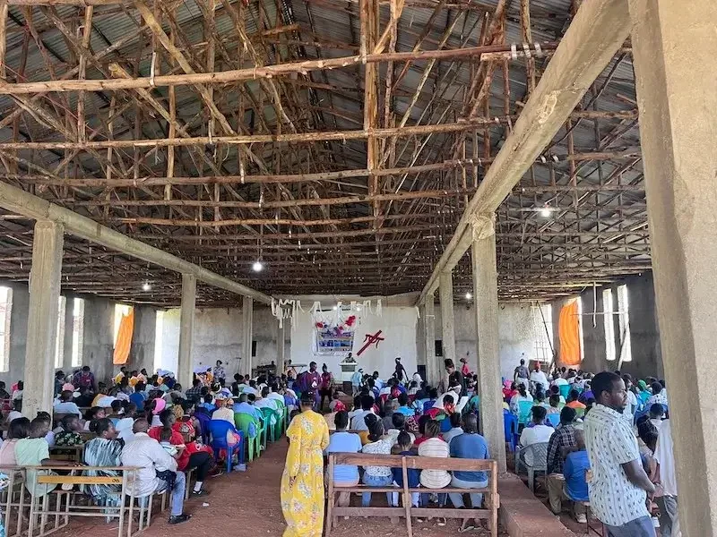 A large gathering of people inside a building with a high, wooden beam ceiling, concrete pillars, and open windows. Many seated on plastic chairs facing a stage with a banner and some decorations.