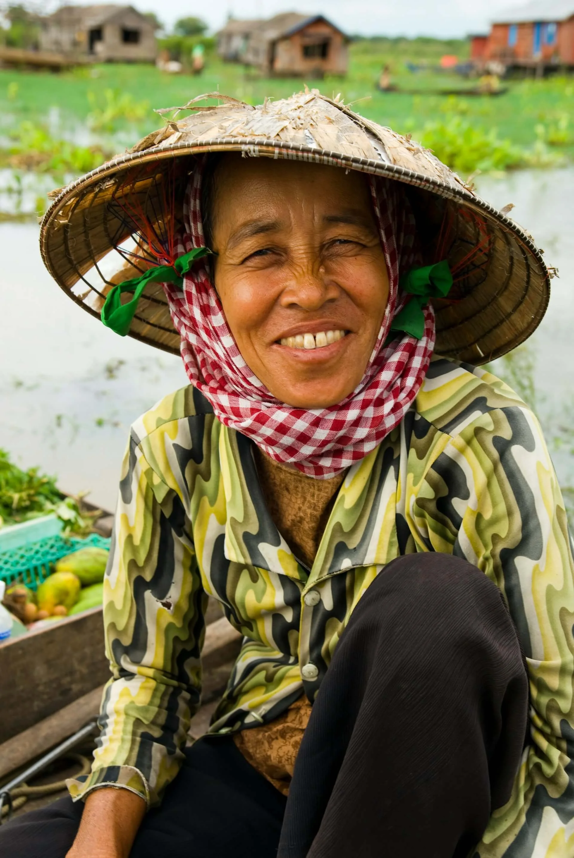 Smiling woman wearing a straw hat with a red checkered scarf around her neck, sitting in a boat near a flooded area with small houses in the background.