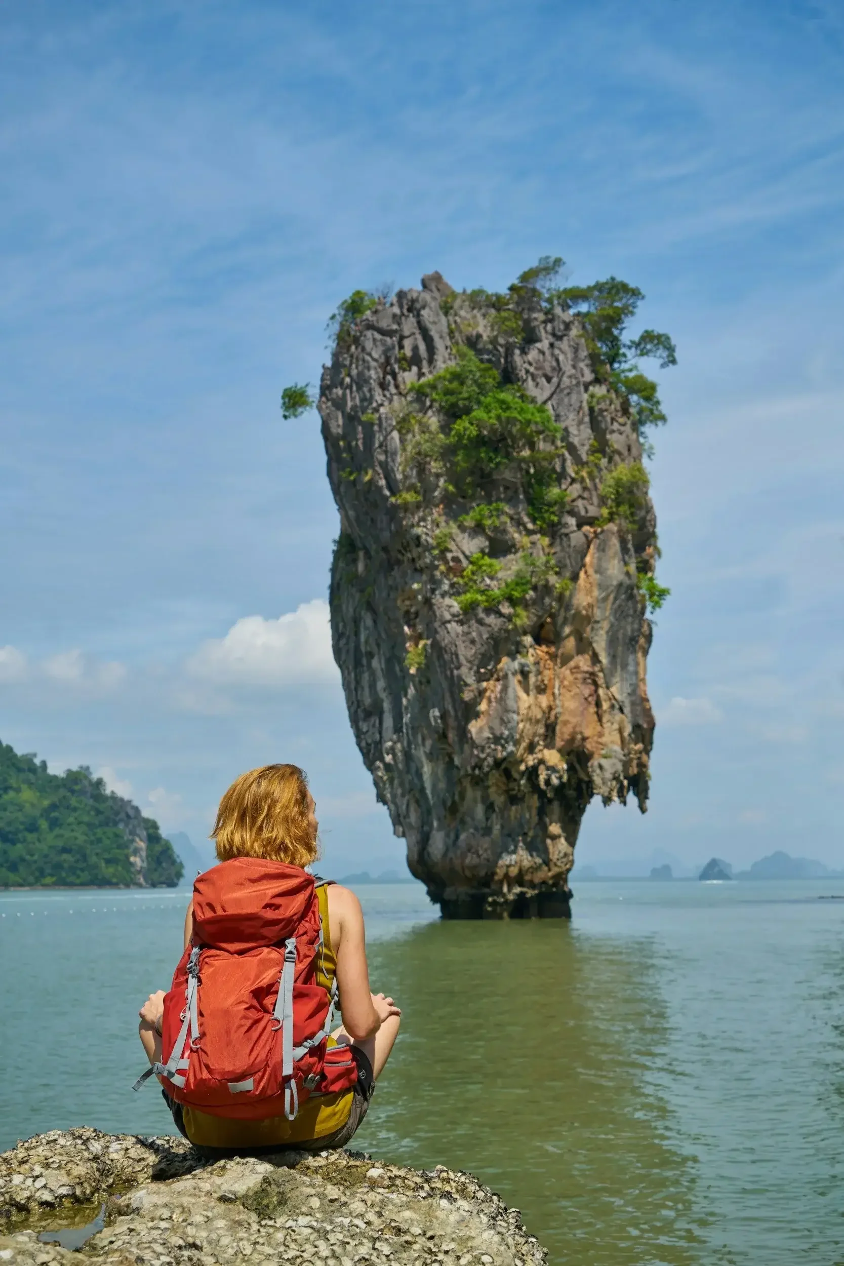 A woman with a red backpack and yellow top sitting on rocks at a beach, looking at a tall, rocky island formation with greenery in the background under a partly cloudy sky.