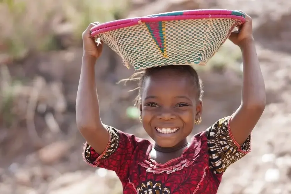 Smiling young girl with earrings balancing a woven basket on her head outdoors.