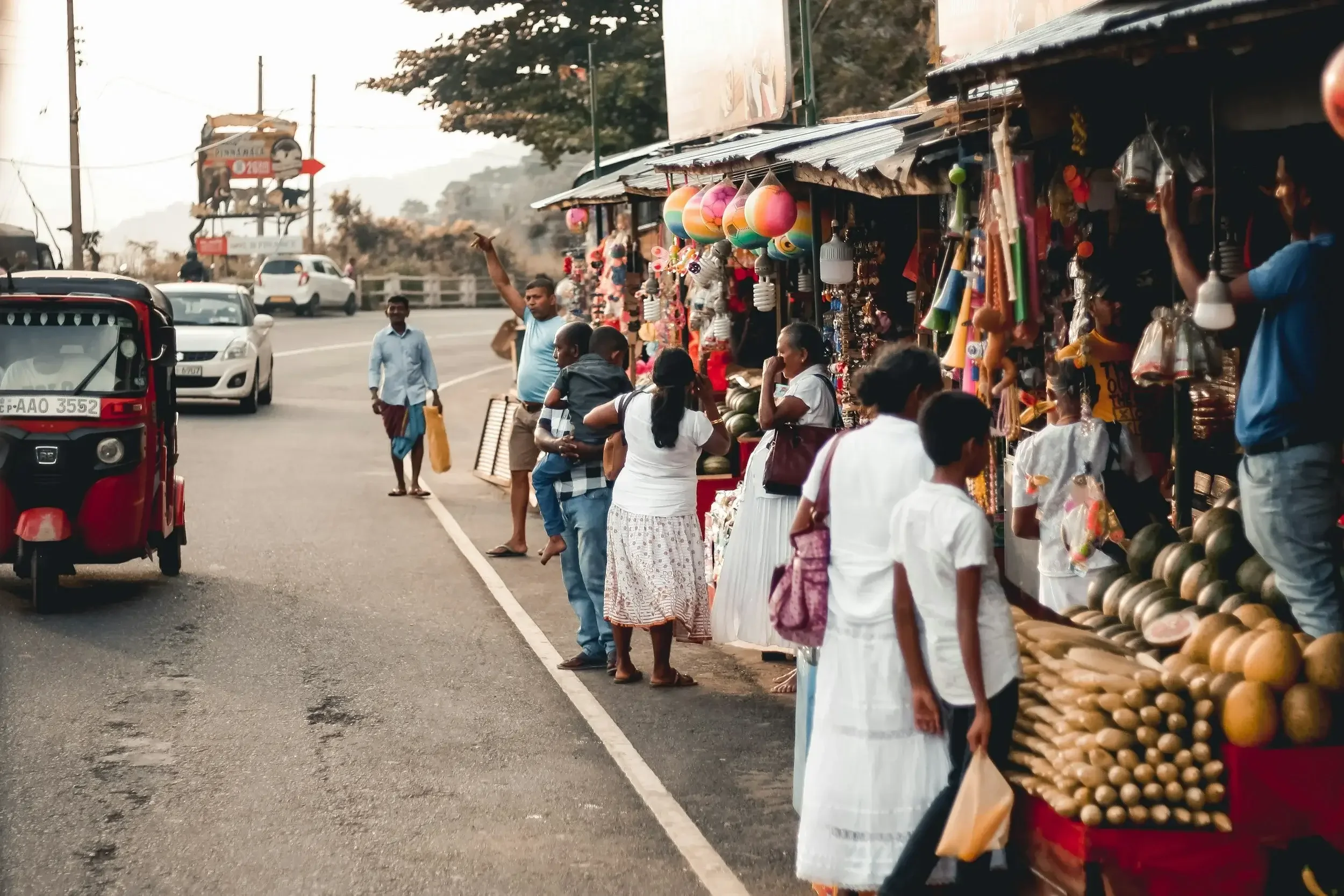 Street scene with a roadside market in a rural area, featuring vendors selling colorful items and people shopping, with vehicles passing by on the road.