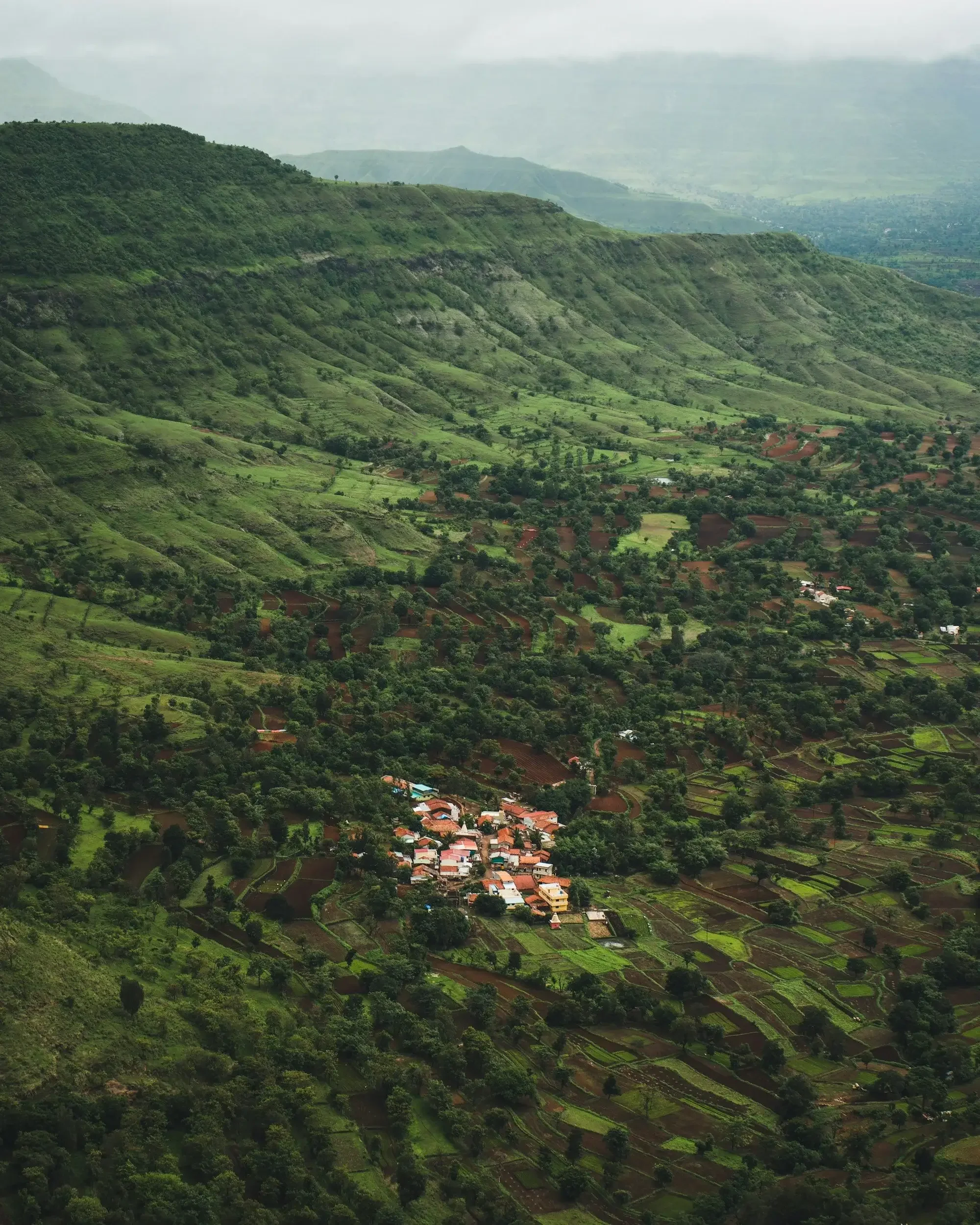 Aerial view of lush green mountains, small village with red roofs, and patchwork farmland.