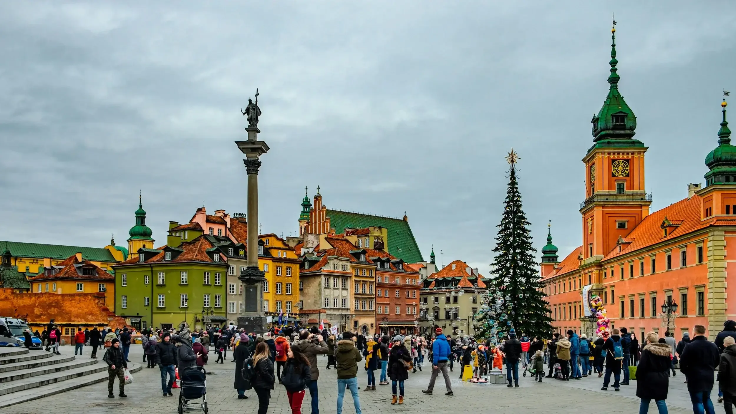 Crowd gathering in a city square with colorful historic buildings, a tall statue on a column, a decorated Christmas tree, and market stalls.