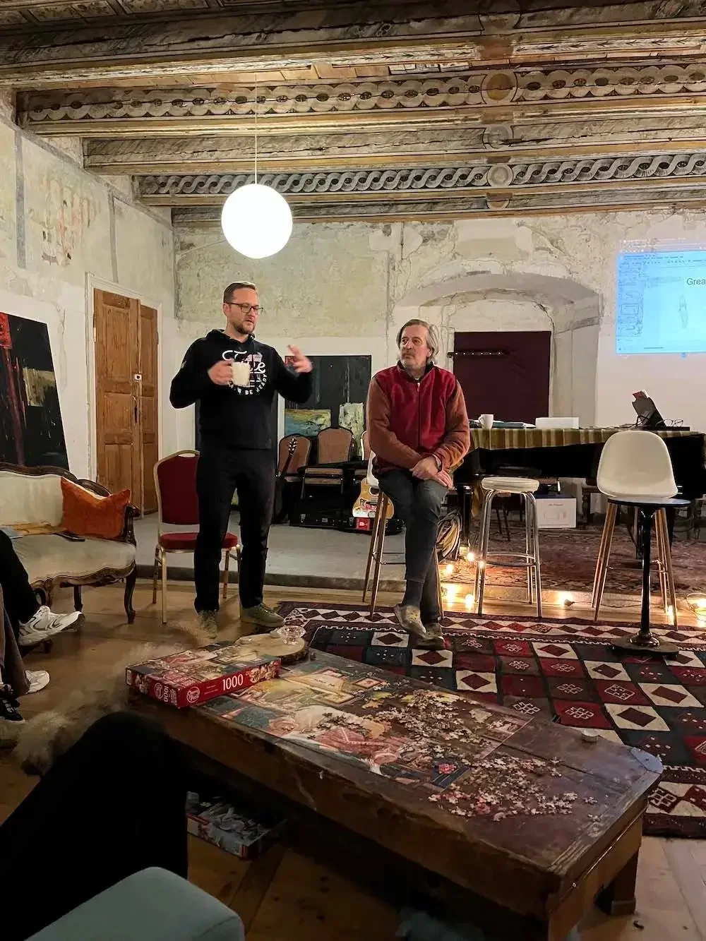 Two men are in a rustic room, one standing and speaking, holding a mug, and the other sitting on a stool. The room has exposed wooden beams and a projection screen. There are chairs, a couch, and a wooden table with a jigsaw puzzle partially assembled on it.