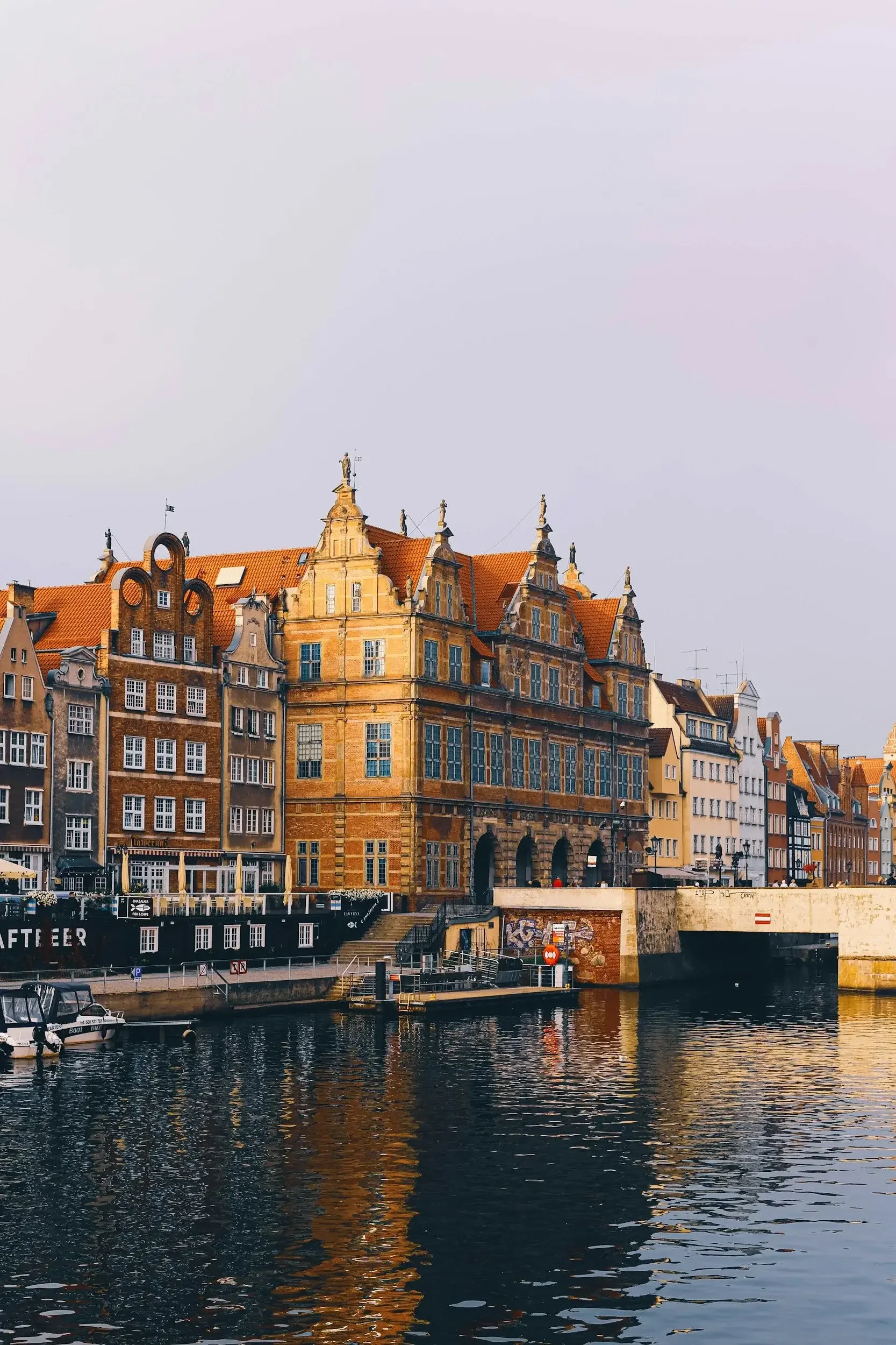 Historic European buildings with red-tiled roofs along a waterfront with boats and reflections in the water.