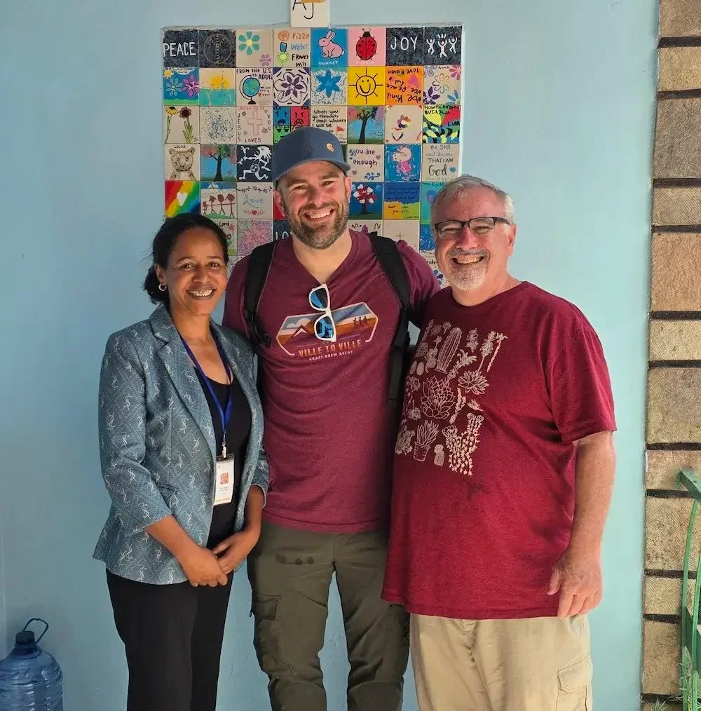 Three people standing together, smiling, in front of a colorful quilt display on a light blue wall. The woman on the left is wearing a gray blazer and black pants, the man in the middle has a backpack, a navy cap, and sunglasses hanging on his shirt, and the man on the right has glasses and a red t-shirt with a white design. A water bottle is on the floor to the left.