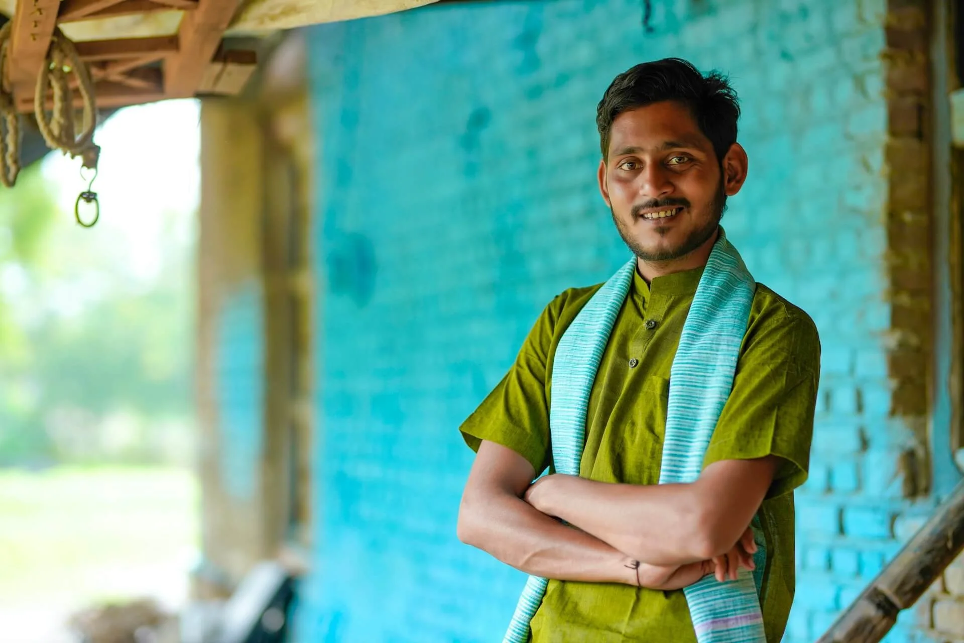 A young man with dark hair and a beard, wearing a green traditional shirt and a light blue scarf, standing outdoors against a blue wall with his arms crossed and smiling at the camera.
