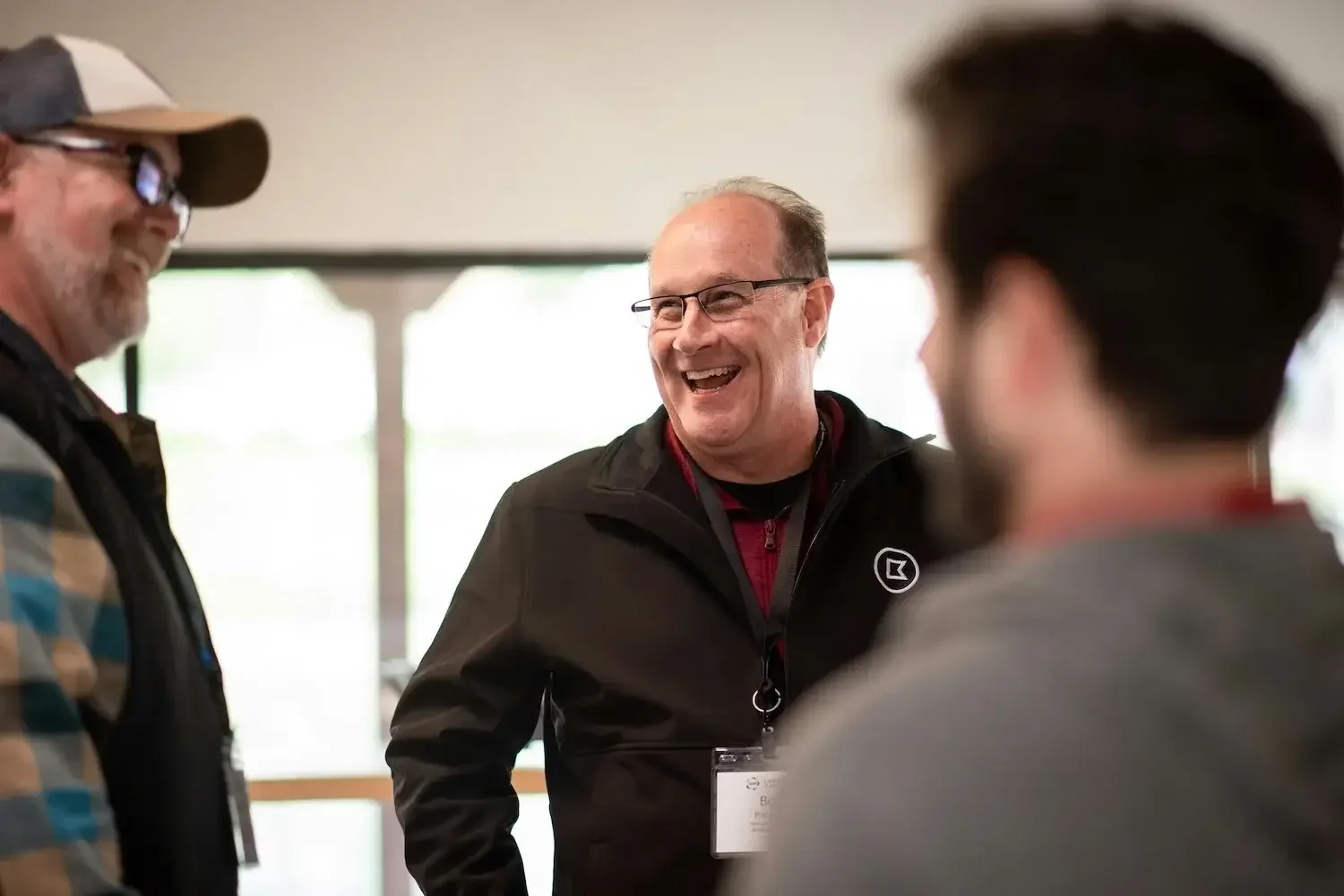 Three men are engaged in a lively conversation indoors, with one man smiling widely and wearing glasses and a black jacket.