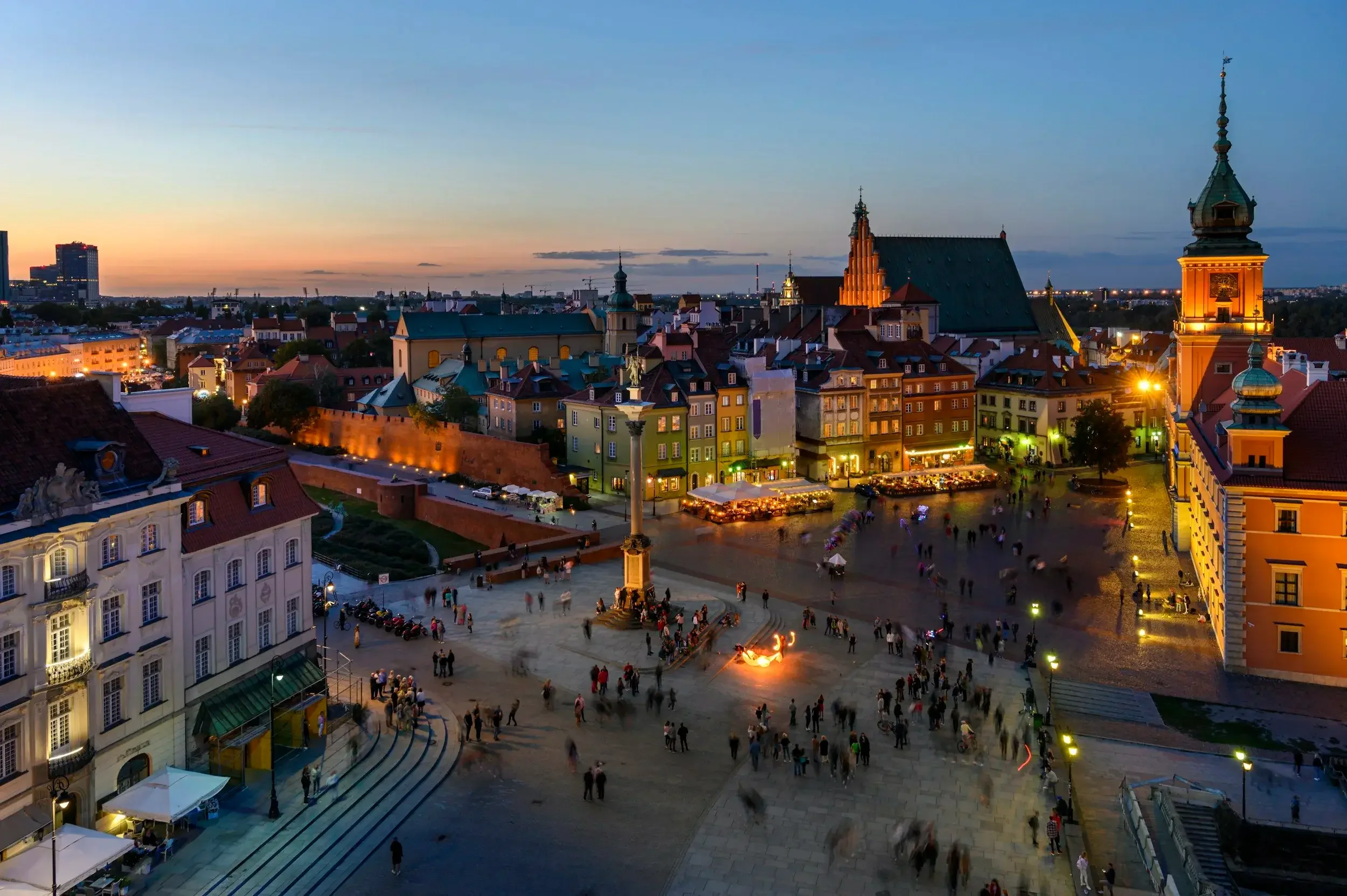 A panoramic view of a historic European city at dusk, featuring a town square with people gathering, illuminated buildings, a church with a tall steeple, and a sunset on the horizon.