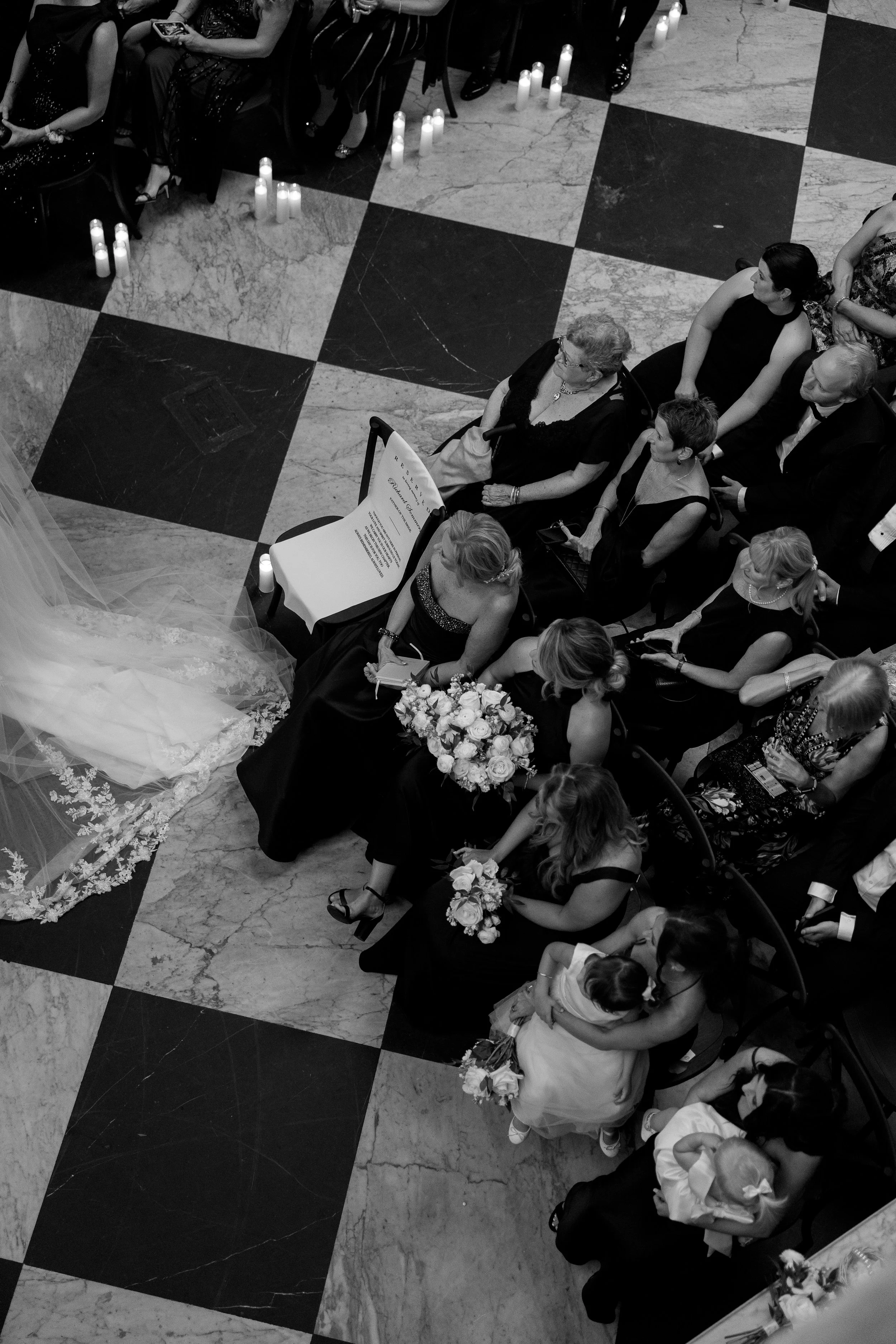 People seated on chairs at a formal event, with candles on the floor along the aisle and a bride holding a bouquet.