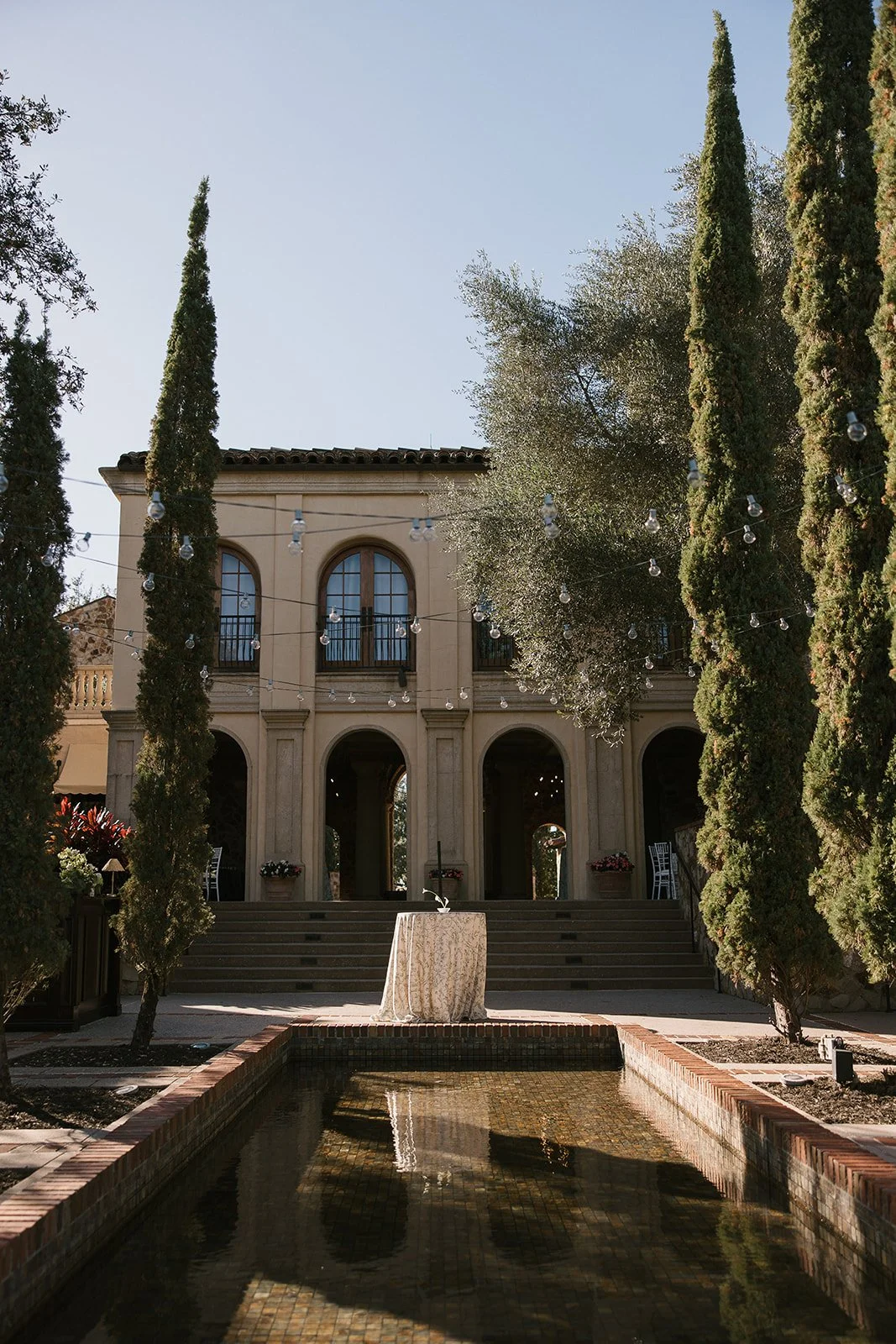 Elegant historic building with arched windows and doorways, surrounded by tall cypress trees, hanging string lights, and a water feature in the foreground.