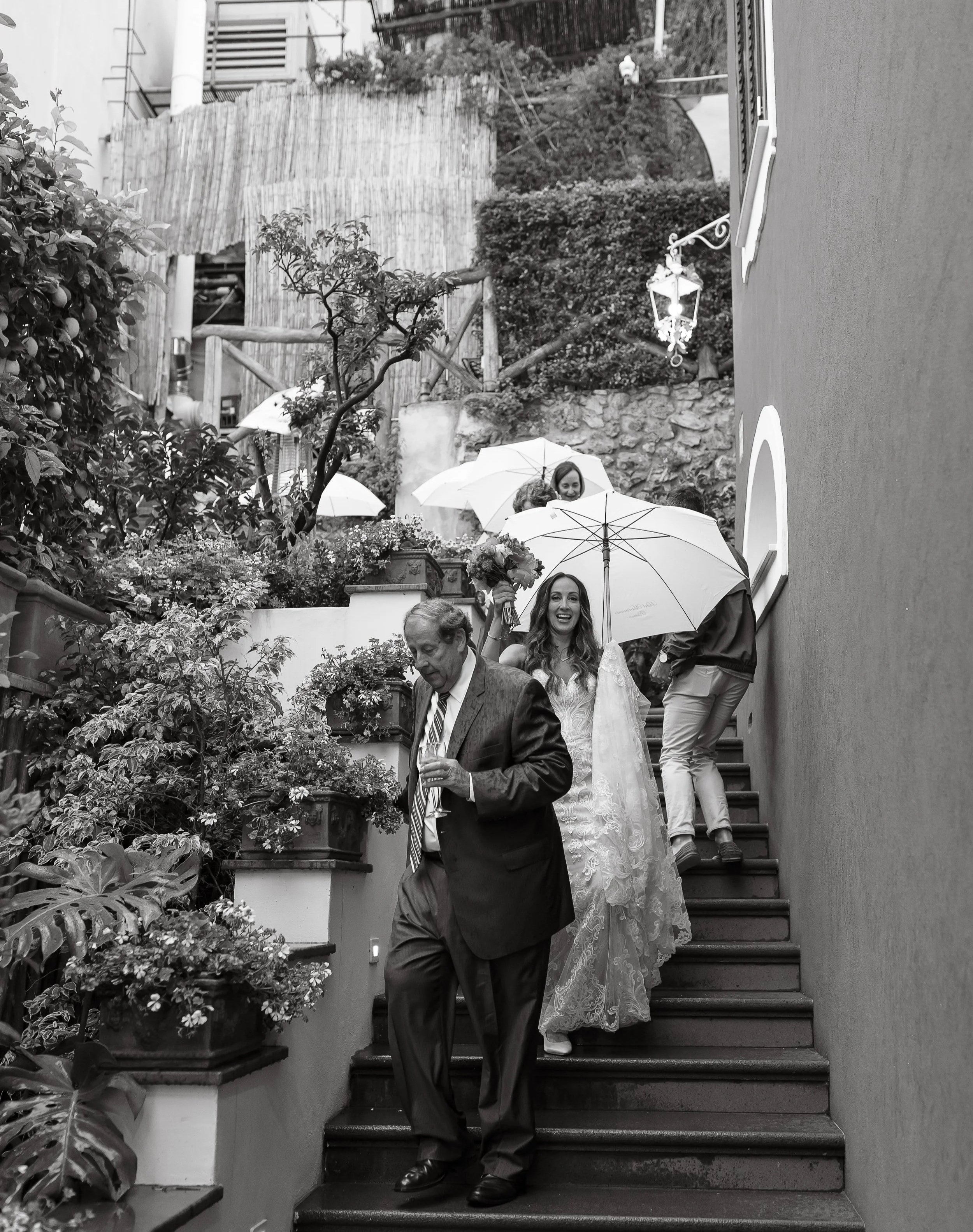 People holding umbrellas walking down outdoor stairs, with lush plants and a stone wall in the background, during a rainy day.