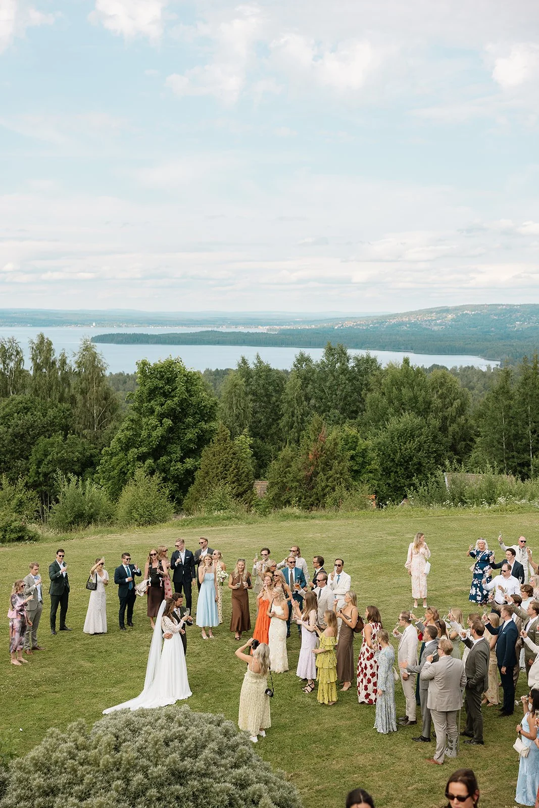 Wedding ceremony outdoors on a grassy lawn near a lake, with many guests dressed in formal attire, trees, hills, and water in the background.