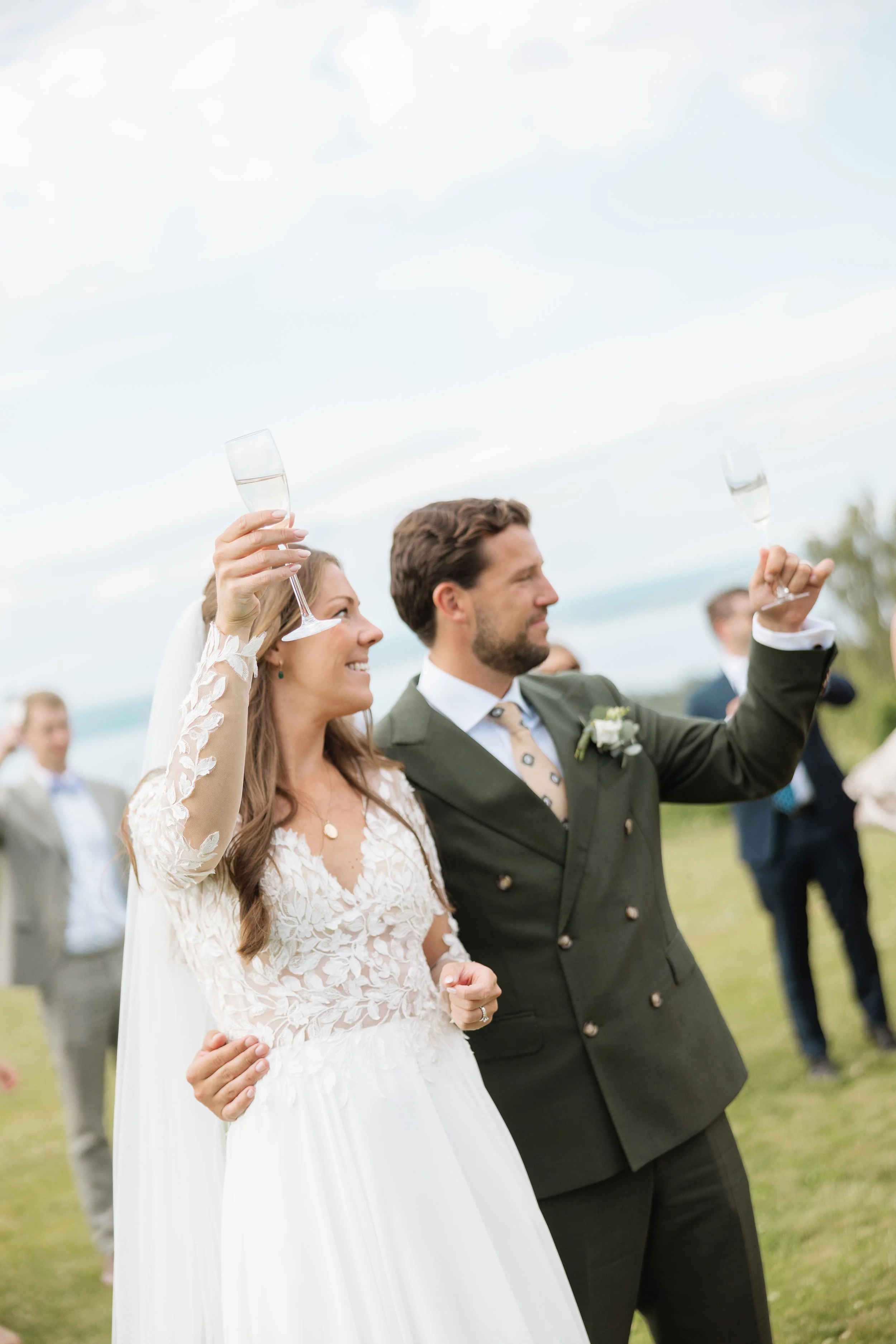 A bride and groom raising champagne glasses during their outdoor wedding celebration, with wedding guests visible in the background.