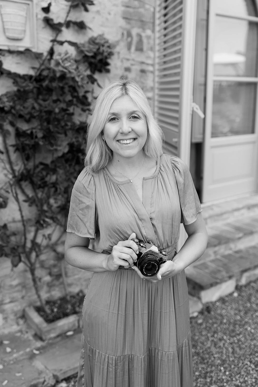 A smiling woman with shoulder-length blonde hair, holding a camera, standing outdoors in front of a stone wall with plants and a window with shutters.