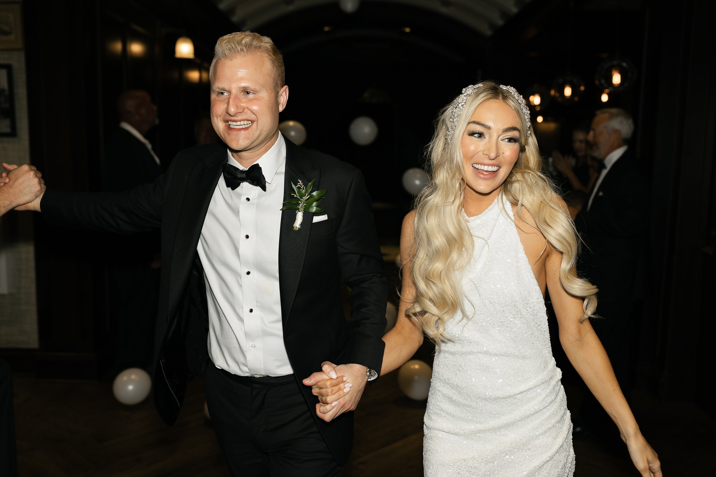 A newlywed couple holding hands and smiling at a wedding reception. The groom wearing a black tuxedo and the bride in a white lace wedding dress with a beaded headband.