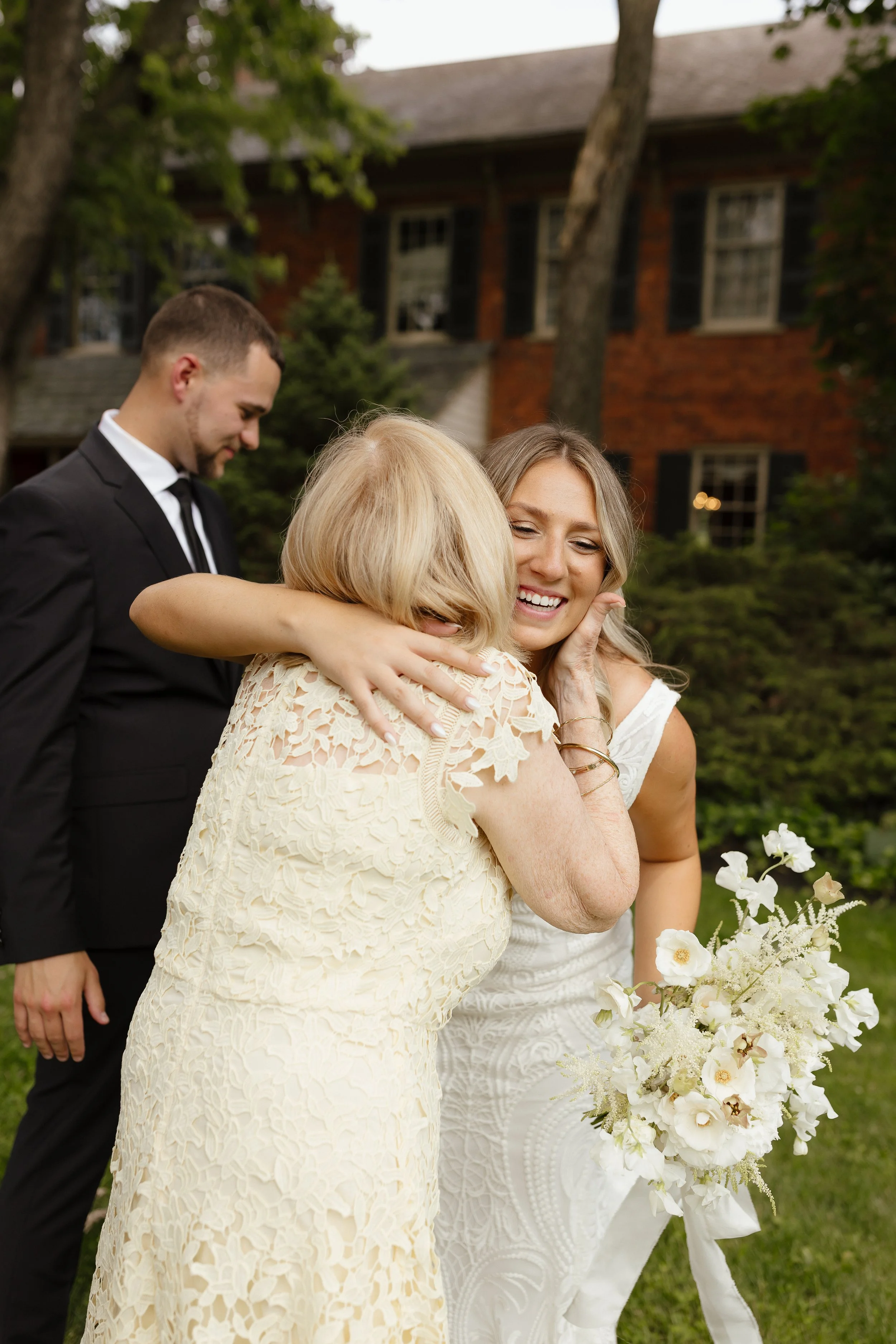 A girl in a white dress hugging an older woman in a cream-colored lace dress, with a bouquet of white flowers, during an outdoor gathering. A young man in a black suit stands in the background.
