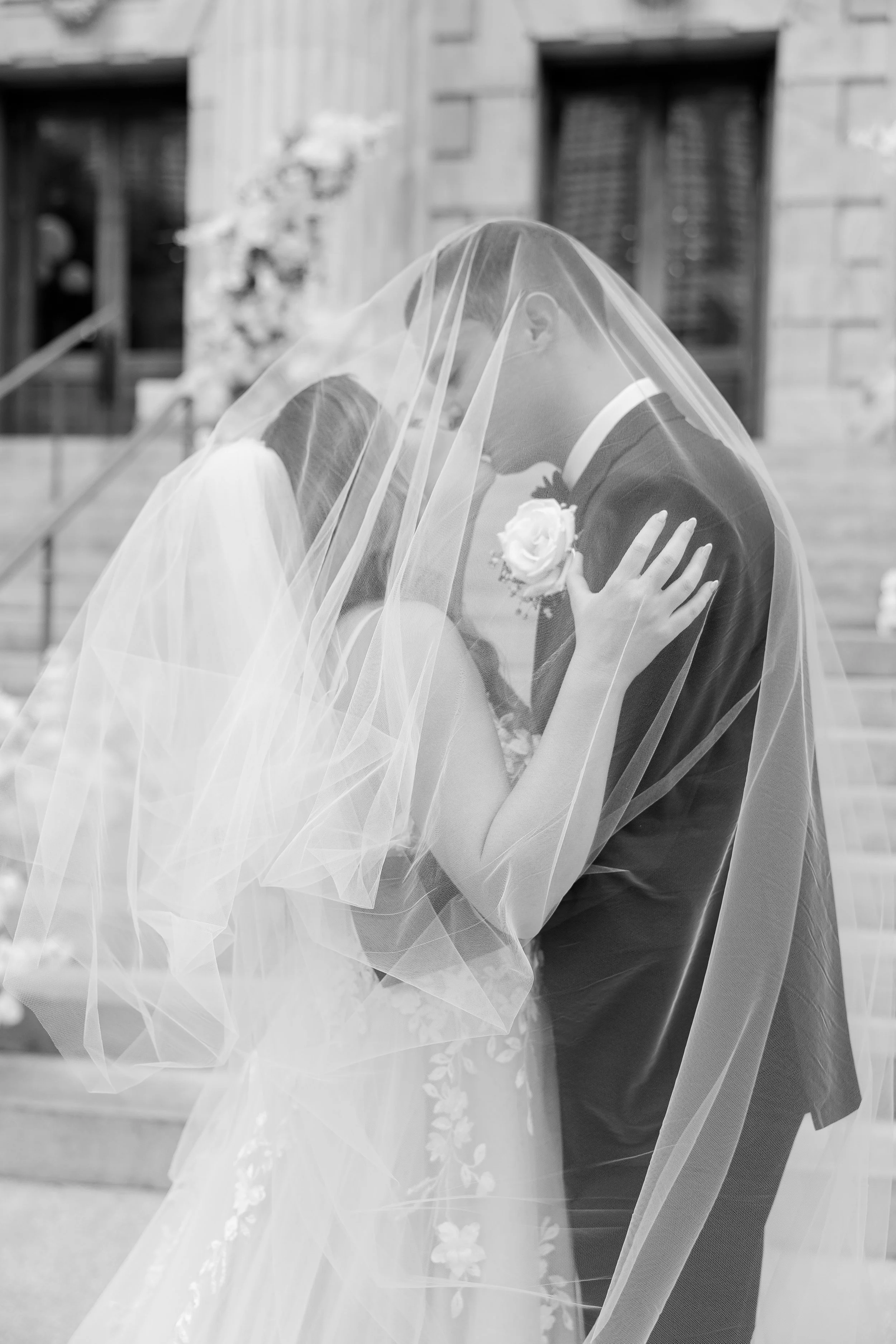 A bride and groom kissing under a sheer veil during a wedding ceremony outside, with a background of buildings and stairs.