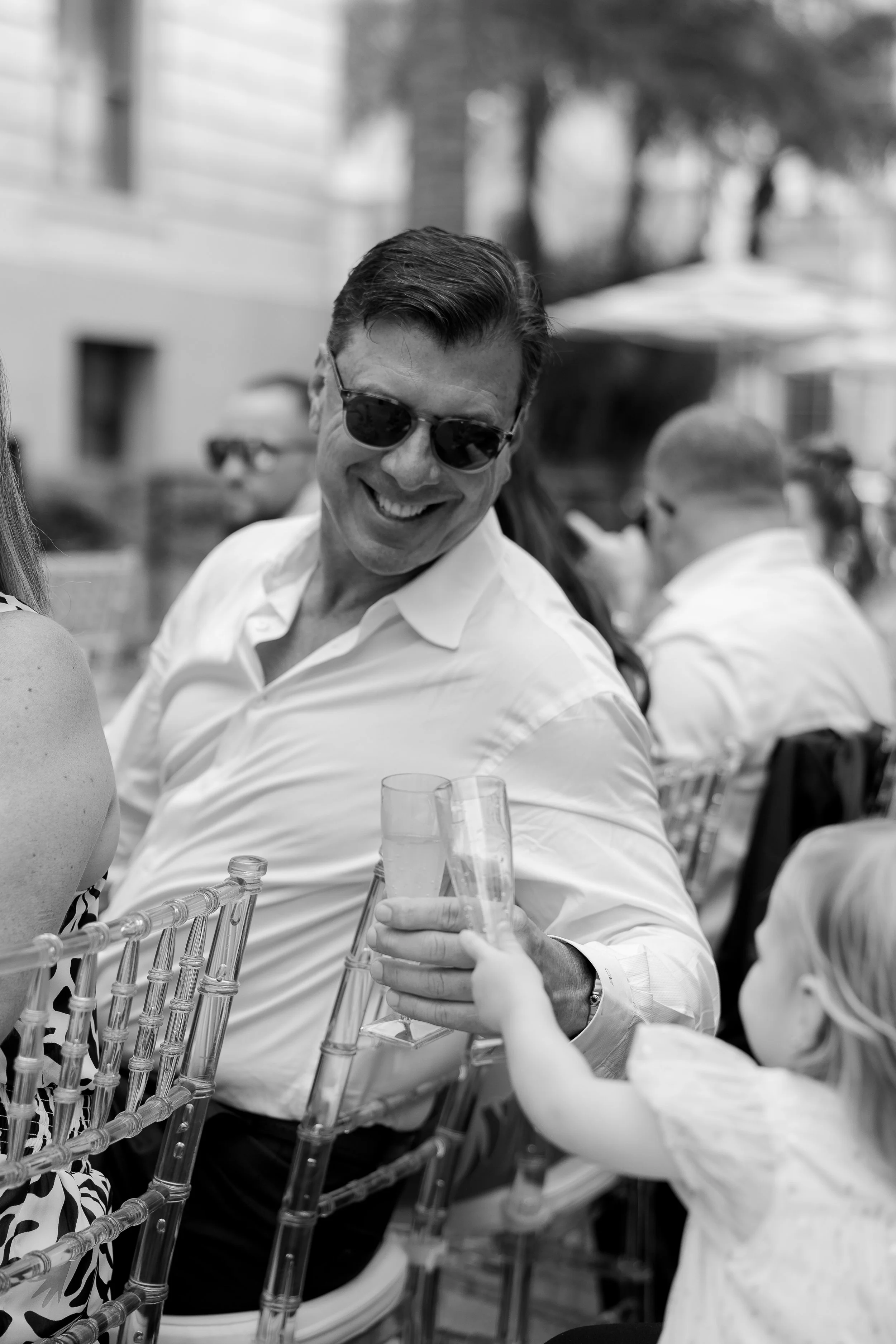 A man wearing sunglasses and a white shirt at a daytime outdoor event, smiling and holding a glass of beverage, surrounded by other people seated at tables.