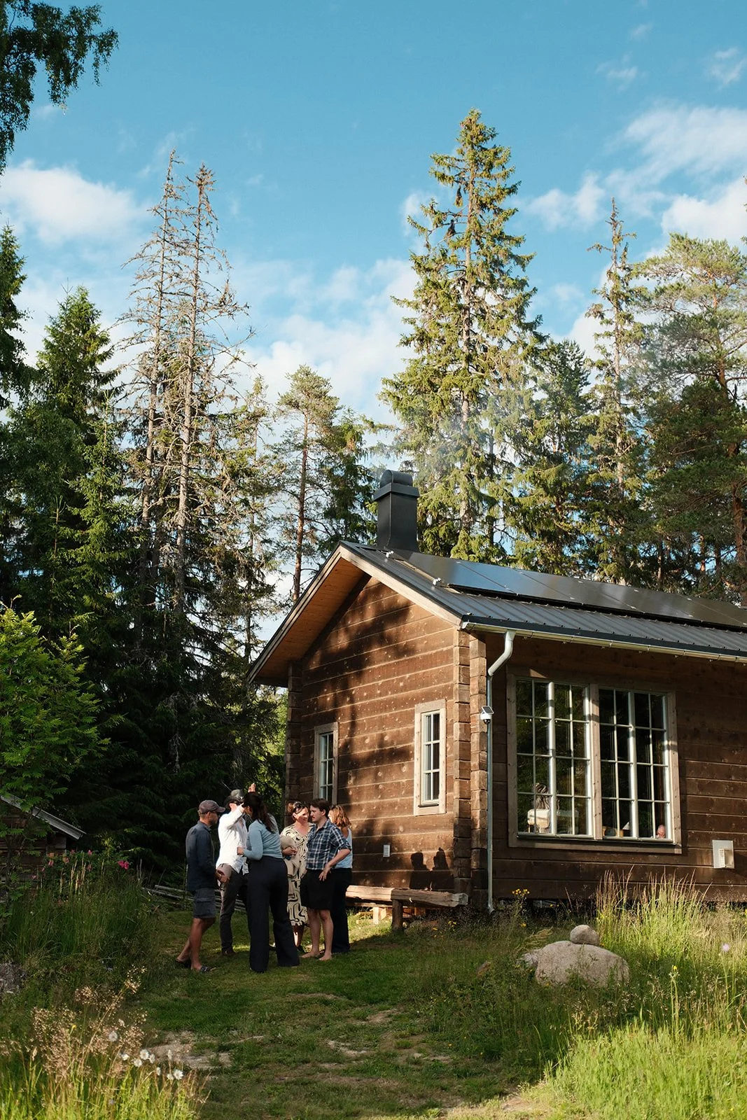 Group of people gathered outside a wooden house in a forested area with tall trees and a blue sky.