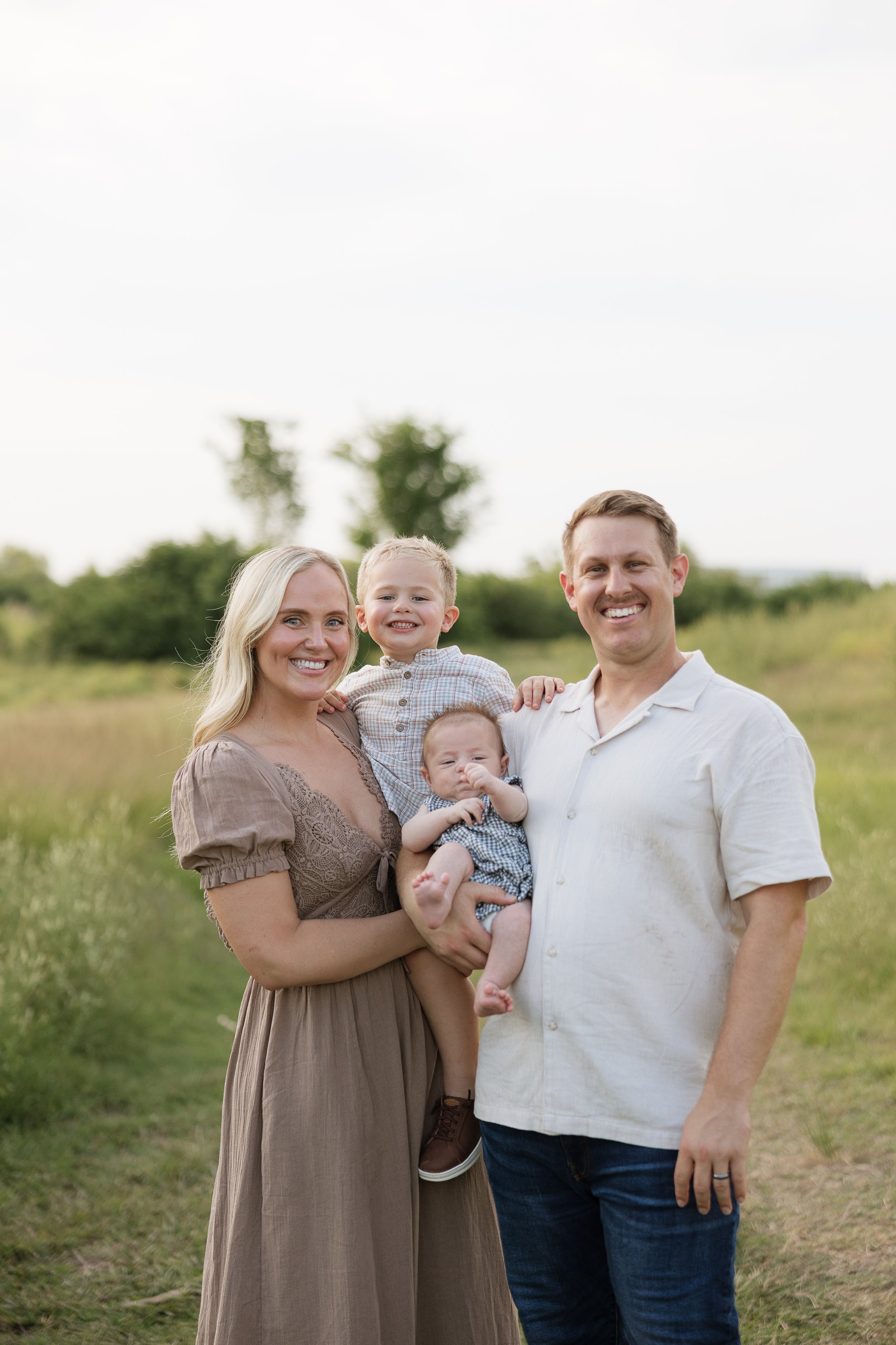 A happy family of four standing outdoors in a grassy field on a cloudy day, smiling at the camera. The family includes a woman, a man, a young boy, and a baby girl.