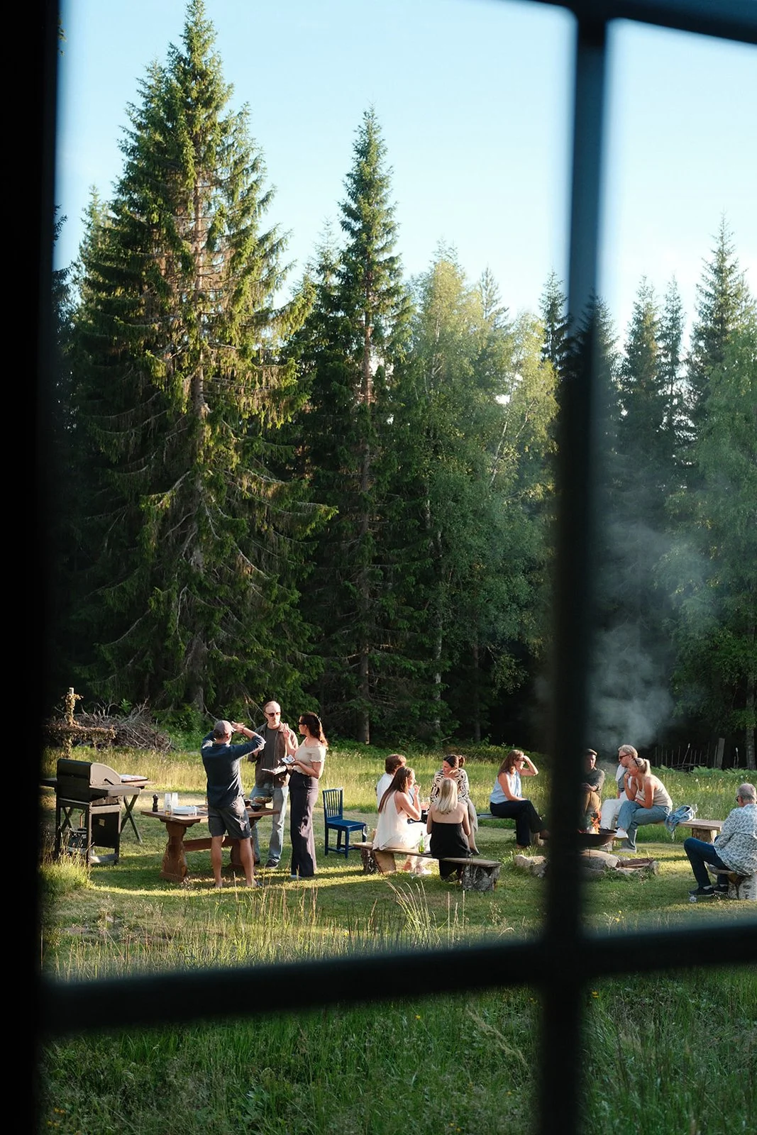 People gathering outdoors in a grassy area with tall pine trees, seen through a window with black framing, with some sitting on benches and others standing, socializing, and a smoker emitting smoke in the background.