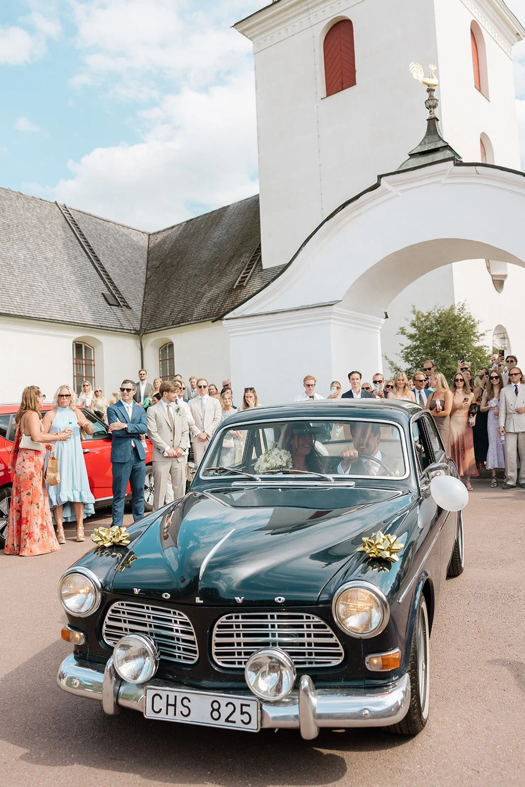 A black classic car decorated with white balloons and gold bows with a bride and groom inside, pulling up outside a white church while guests gather around