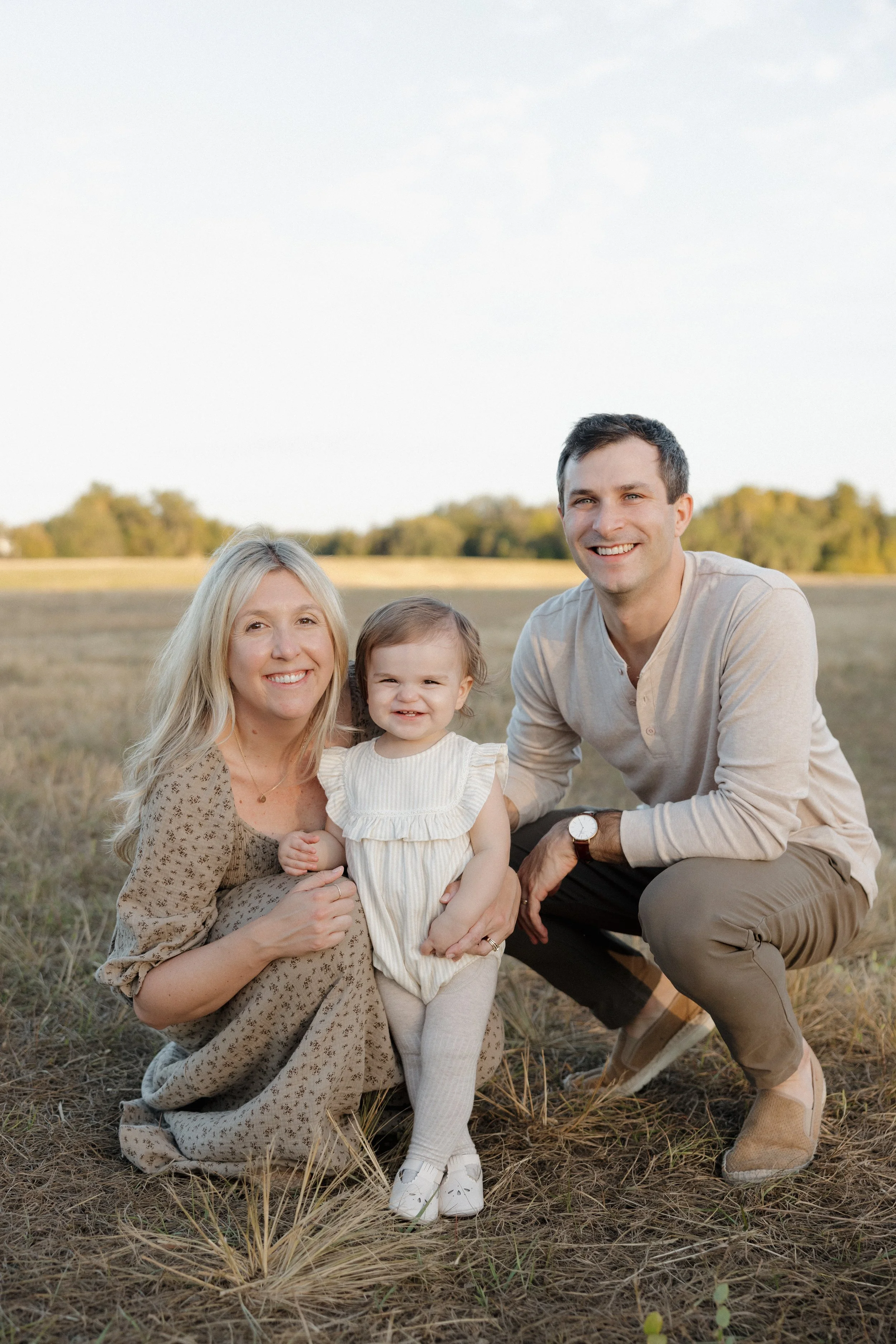 A family of four, including an older woman, a young girl, a young man, and a woman, smiling and posing outdoors in a field with dry grass and trees in the background, during late afternoon or early evening.