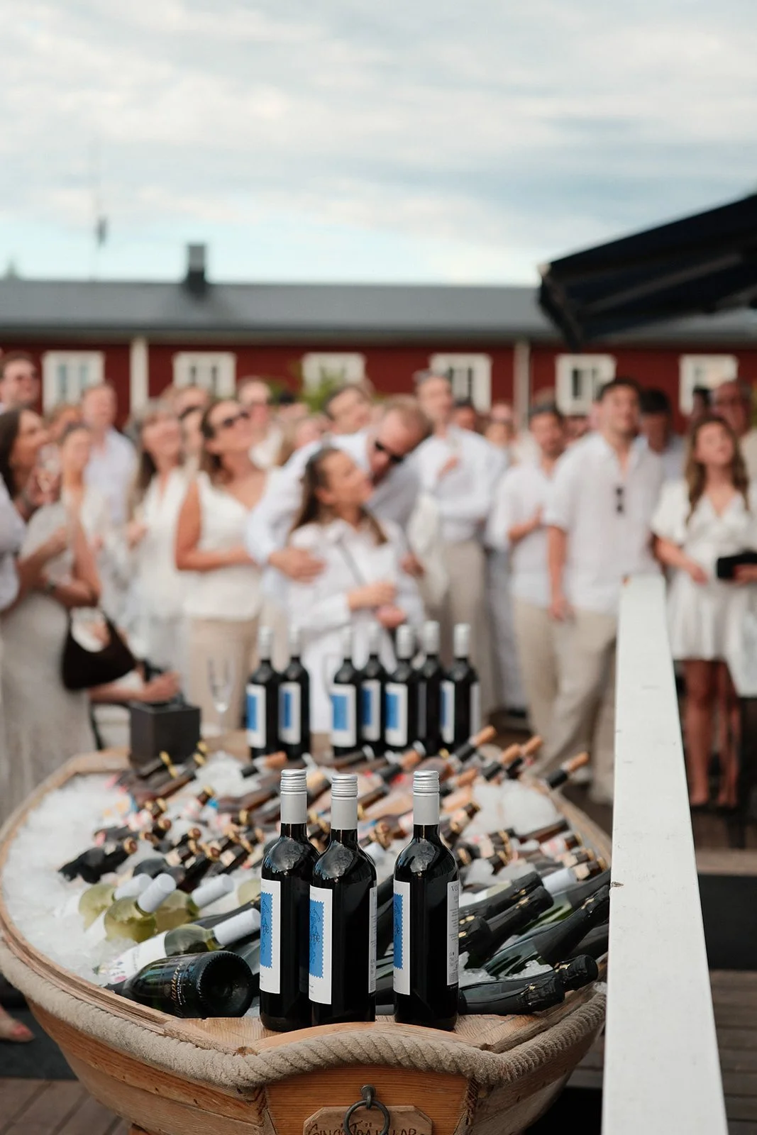 Bottles of wine laid out on ice in a boat at an outdoor gathering with people dressed in white, attending a wedding reception in a backyard with a red house in the background.