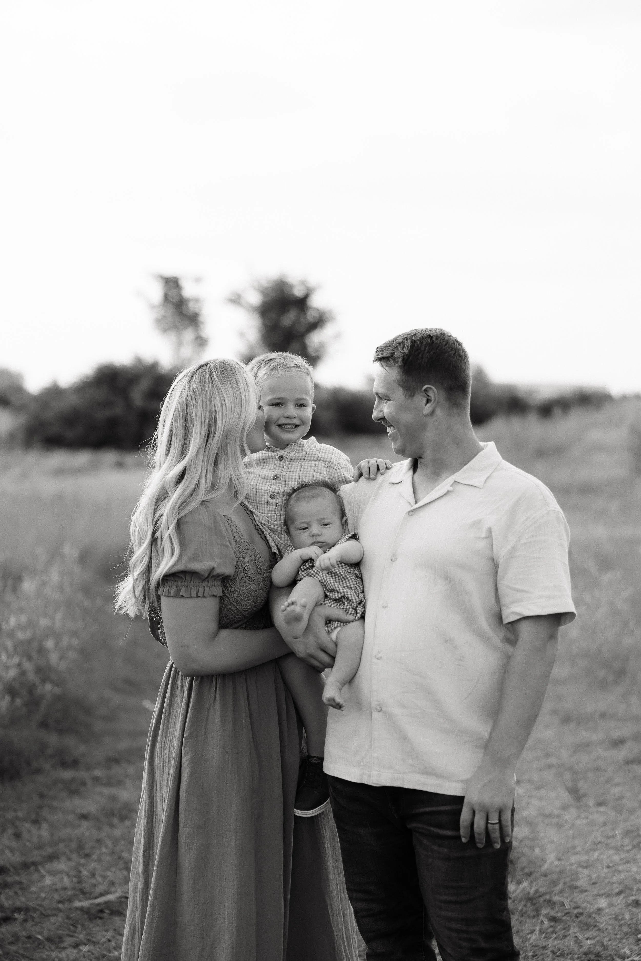 A black-and-white photo of a family of four outdoors, consisting of a woman with long hair, a man, and two young children, one of whom is being held. The family is smiling and close together in a grassy field with trees in the background.