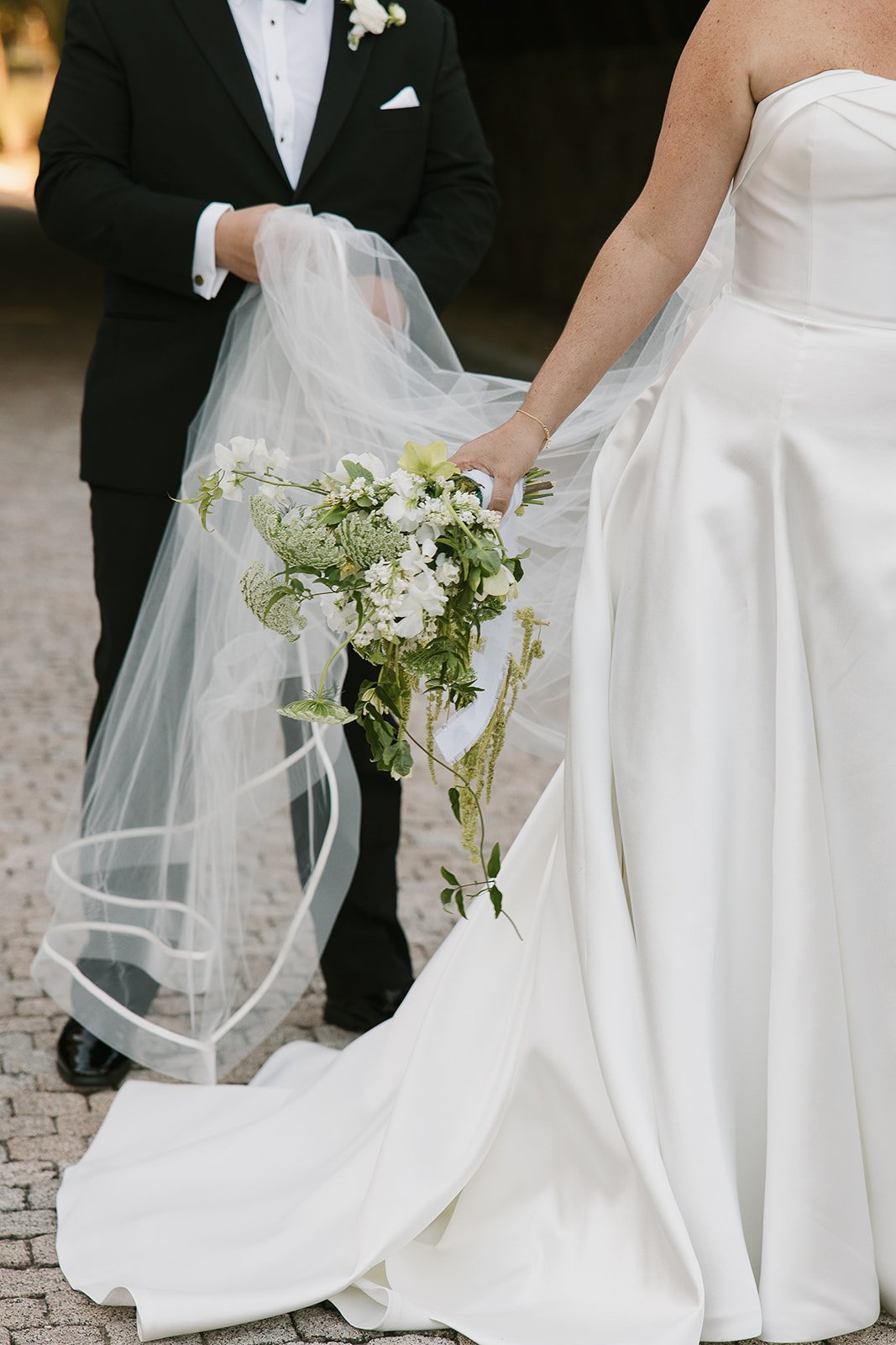 Bride in a white wedding gown holding a bouquet of white and green flowers, standing next to a groom in a black tuxedo, outdoors on a cobblestone path.